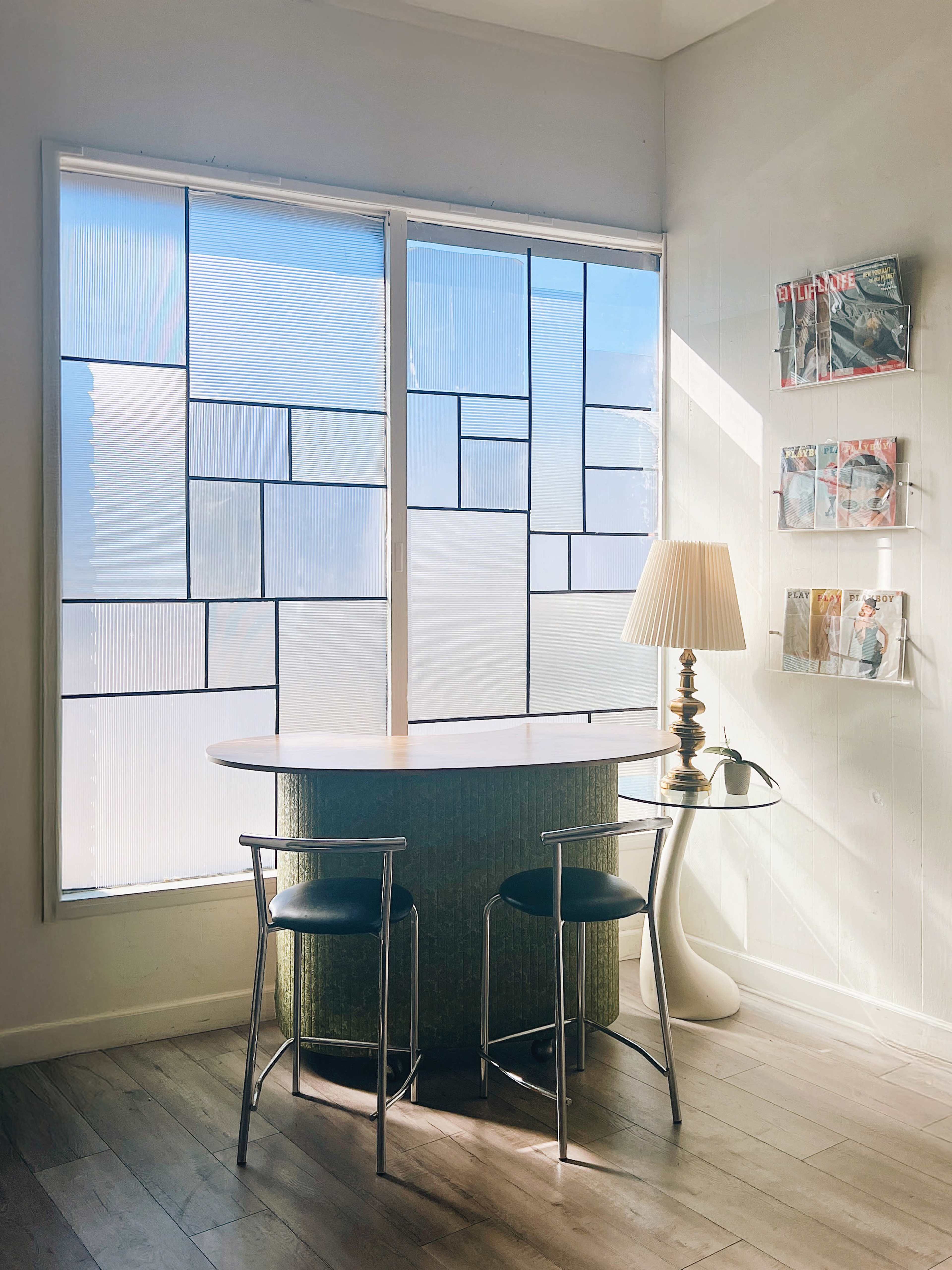A small desk with two chairs is positioned near large windows featuring a geometric design, while a lamp and magazines are displayed nearby.