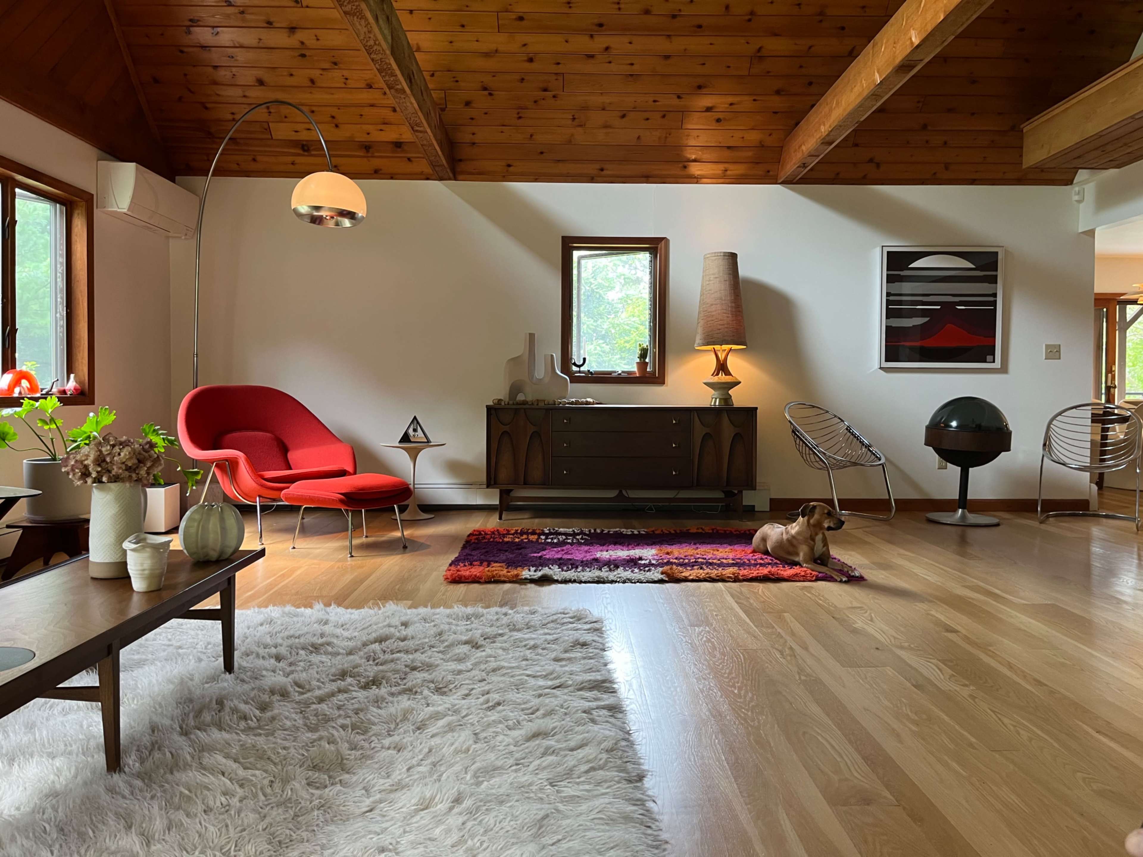 The image shows a spacious living room with wooden beams, a red chair, a coffee table, and a dog resting on a colorful rug.