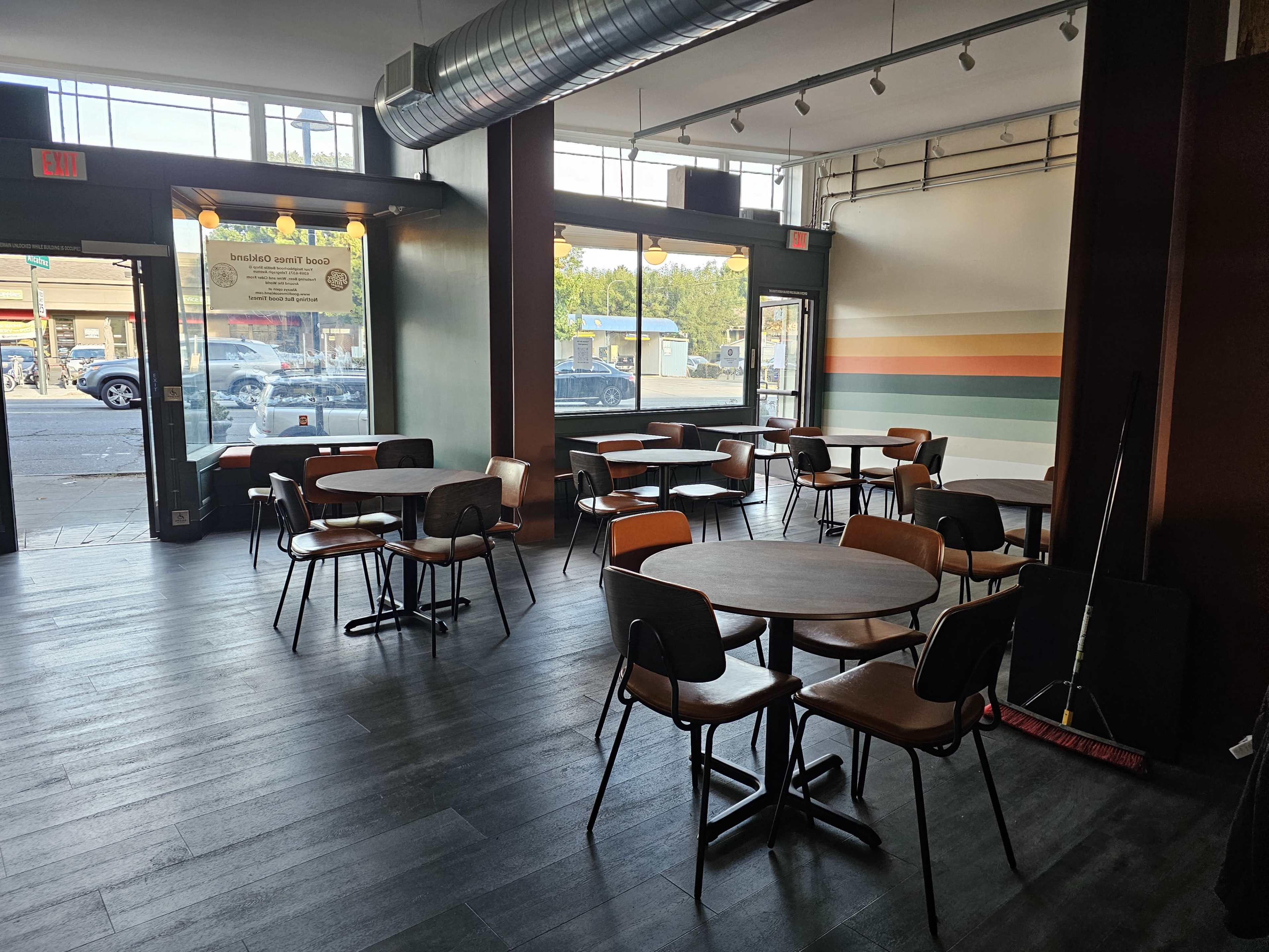 The image shows a spacious, modern café interior with several wooden tables and chairs arranged neatly, large windows allowing natural light, and colorful wall stripes in the background.