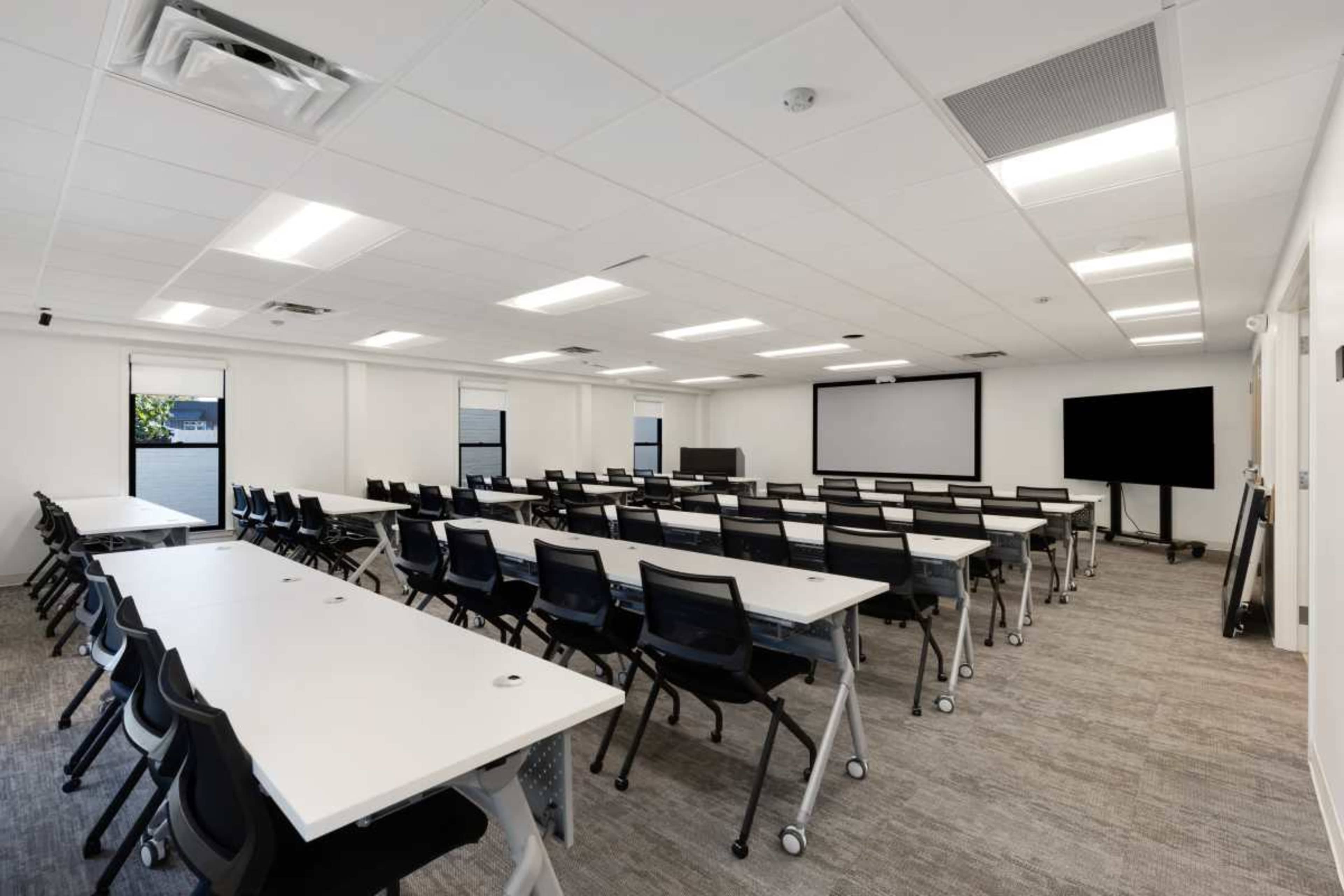 The image shows a modern classroom with rows of white tables and black chairs, equipped with large screens at the front and side walls.