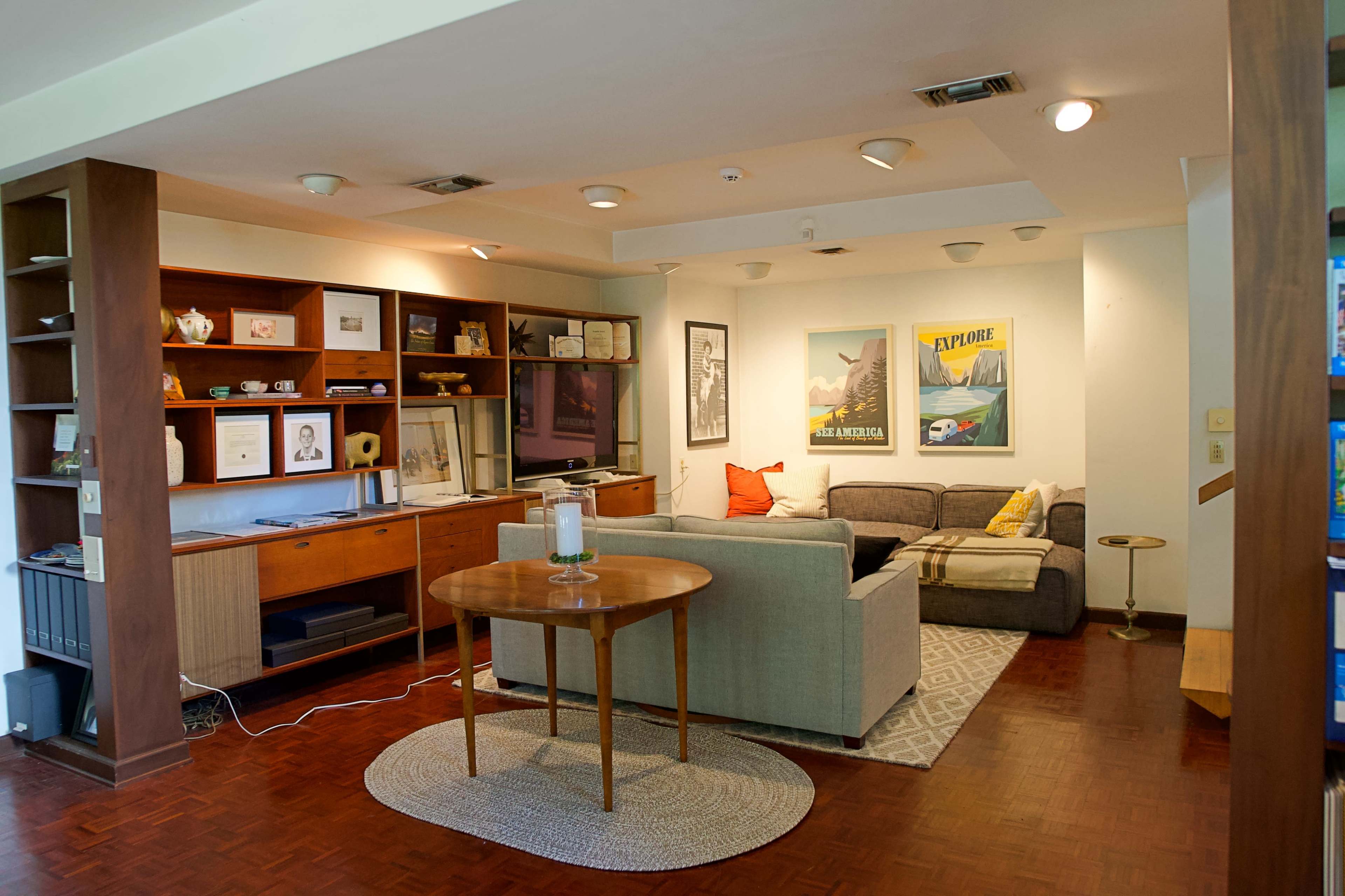 The image shows a modern living room with a couch, coffee table, shelving unit, and various framed artworks on the walls.
