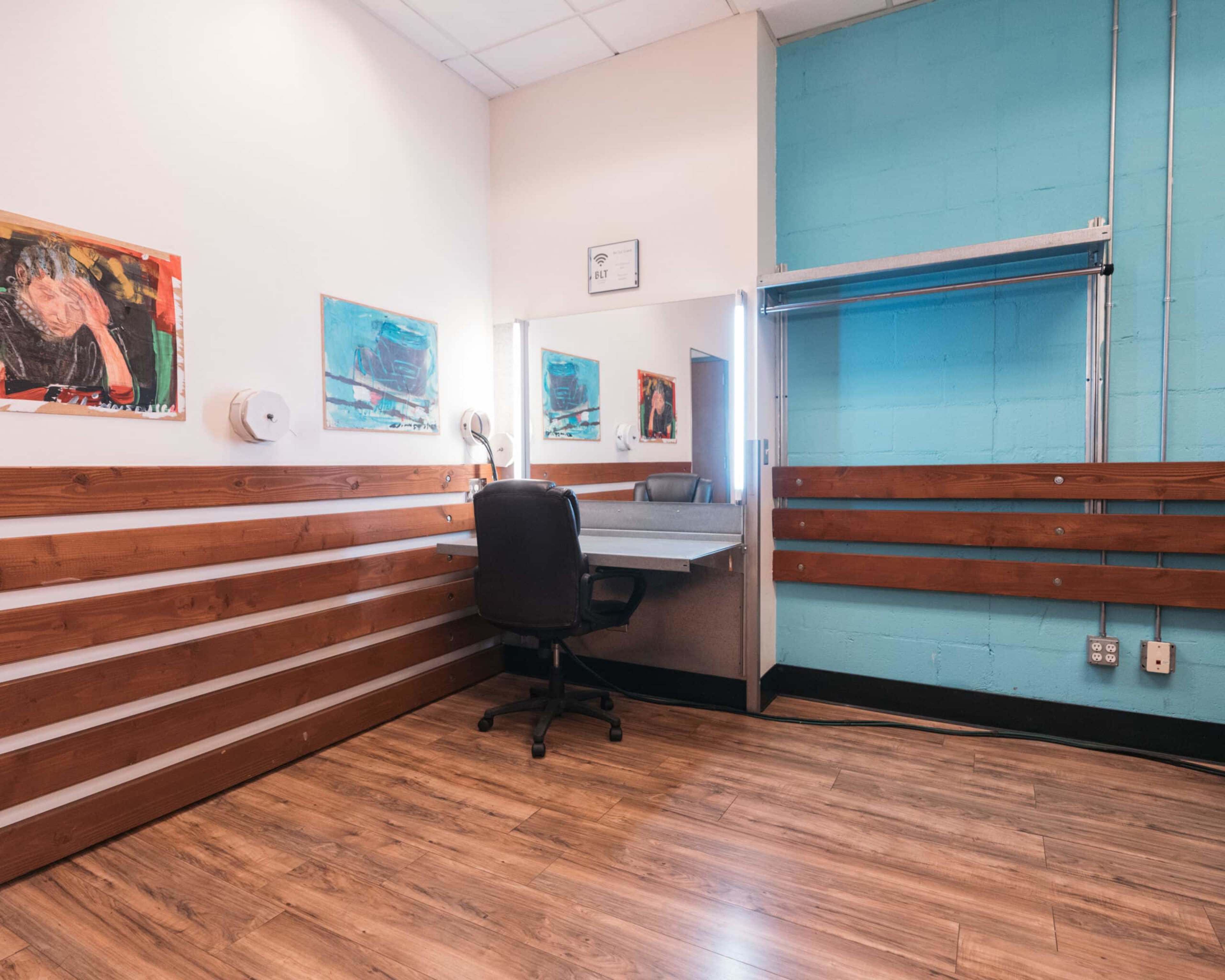 The image shows a small room with a wooden floor, a chair at a dressing table, and colorful paintings on the walls.