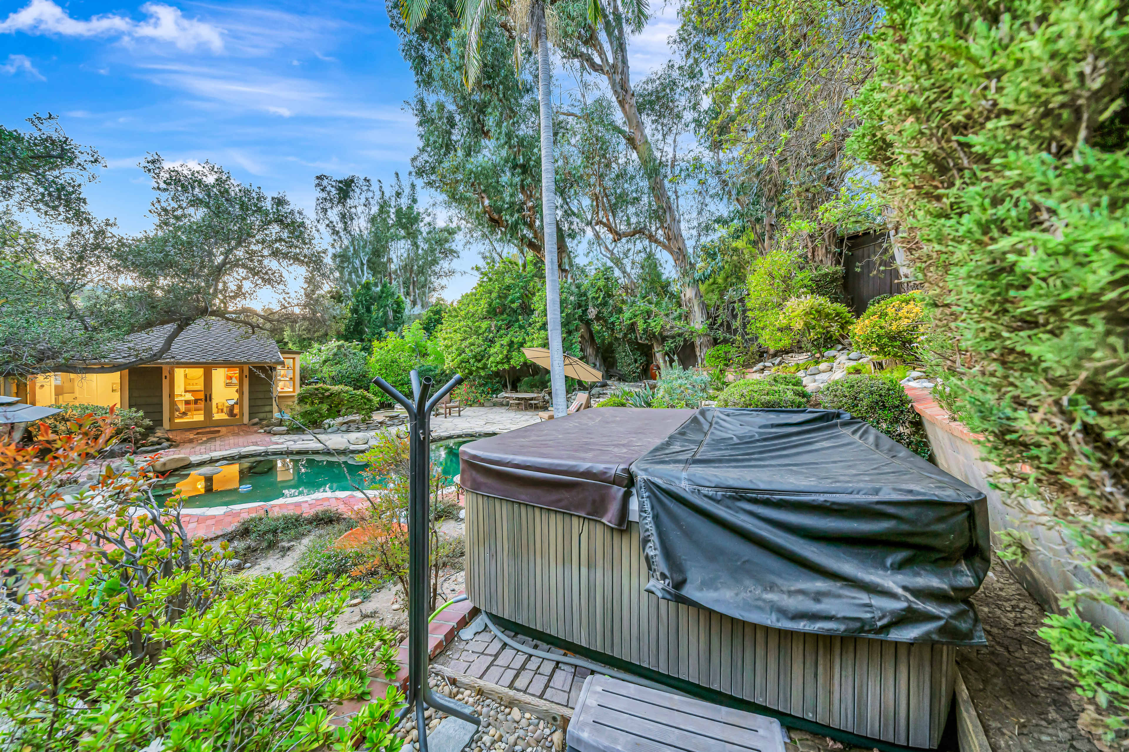 The image shows a secluded backyard featuring a hot tub covered with a black tarp, surrounded by lush greenery and a swimming pool in the background.