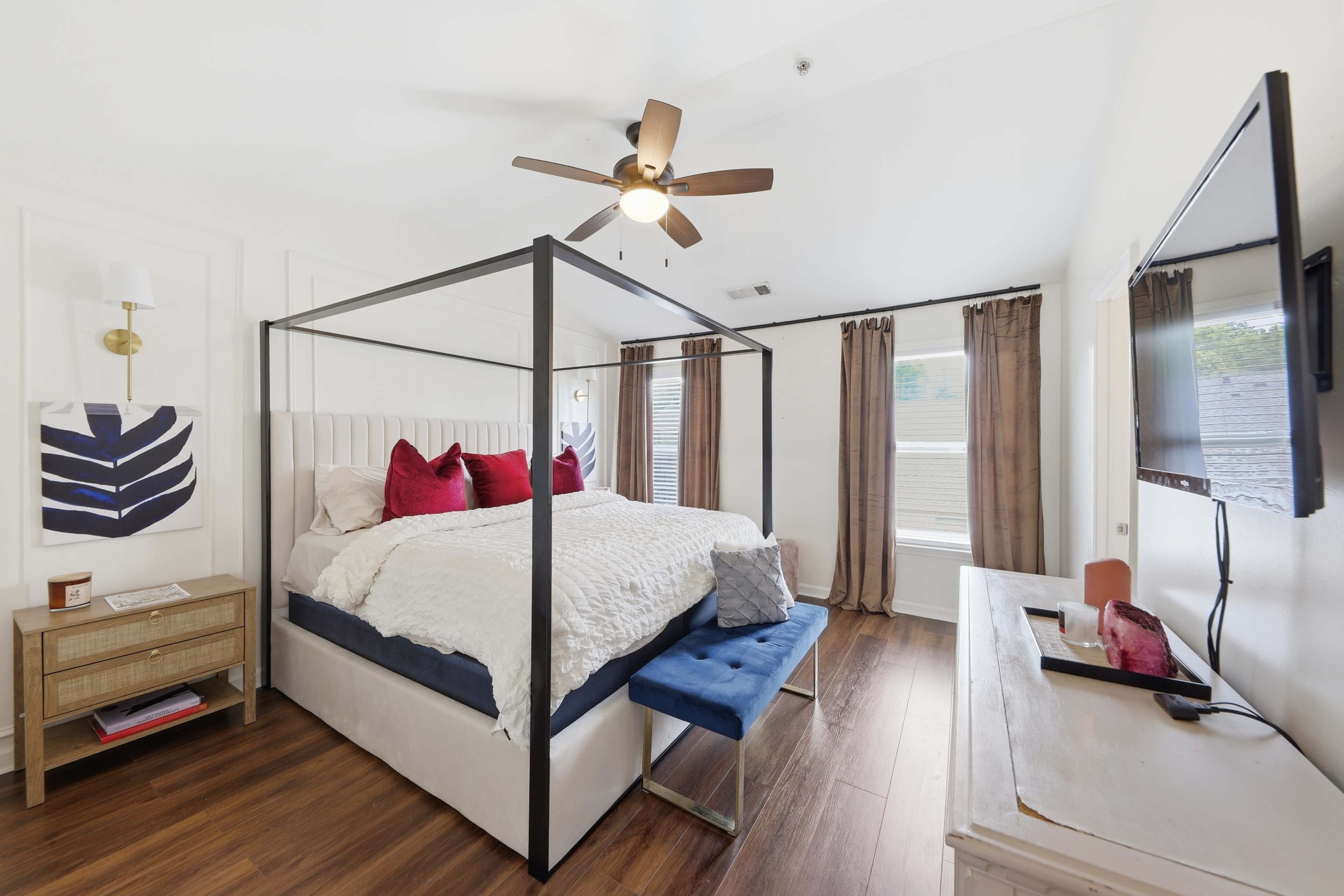 The image shows a well-decorated bedroom featuring a four-poster bed, a sleek bench at the foot, and a wall-mounted television.