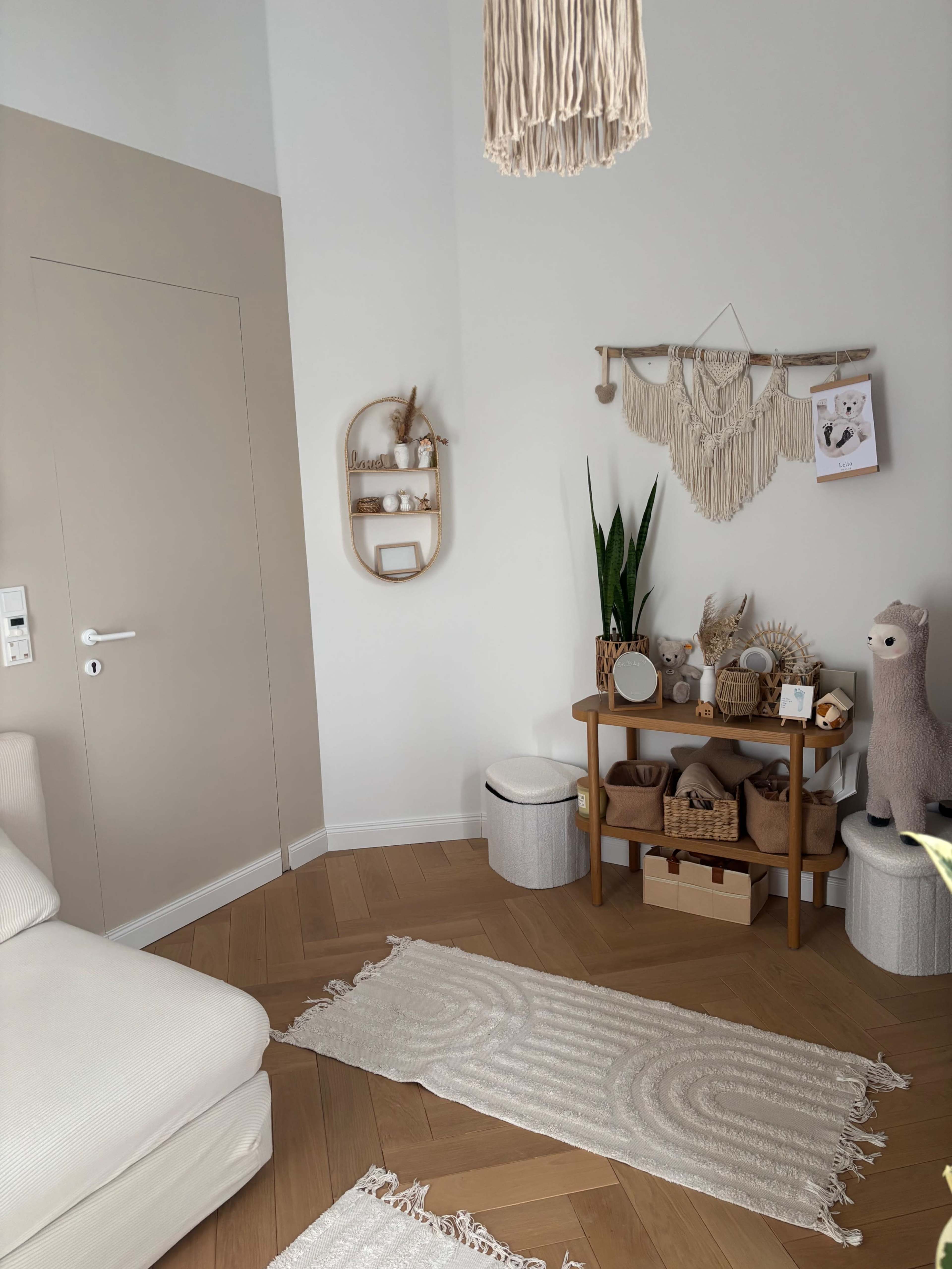 The image shows a cozy and neatly arranged corner of a living room, featuring a beige wall, a white sofa, and a wooden shelf with decorative items and plants.