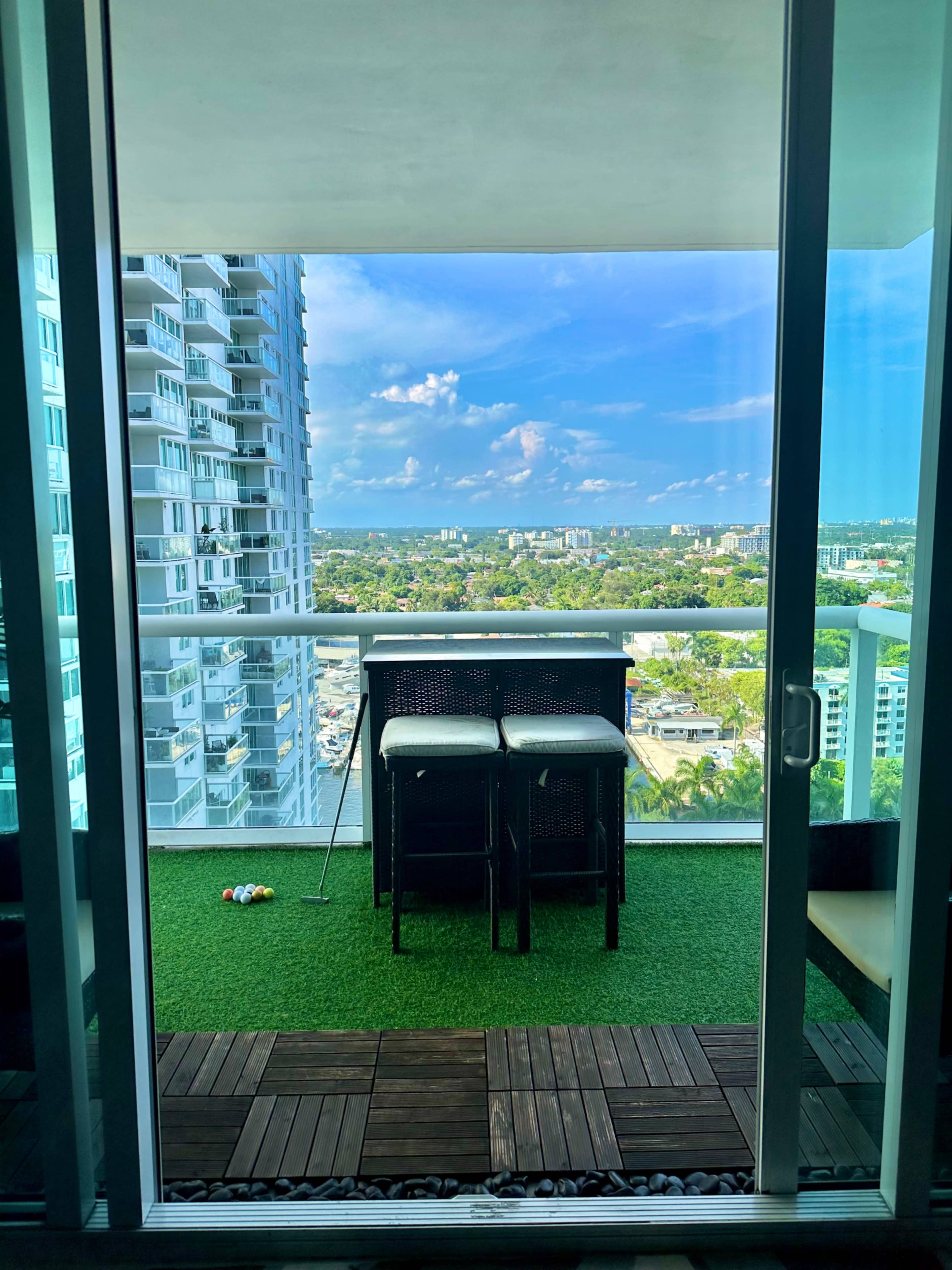 A balcony with a small table and two chairs, overlooking a cityscape with green trees and buildings under a blue sky.
