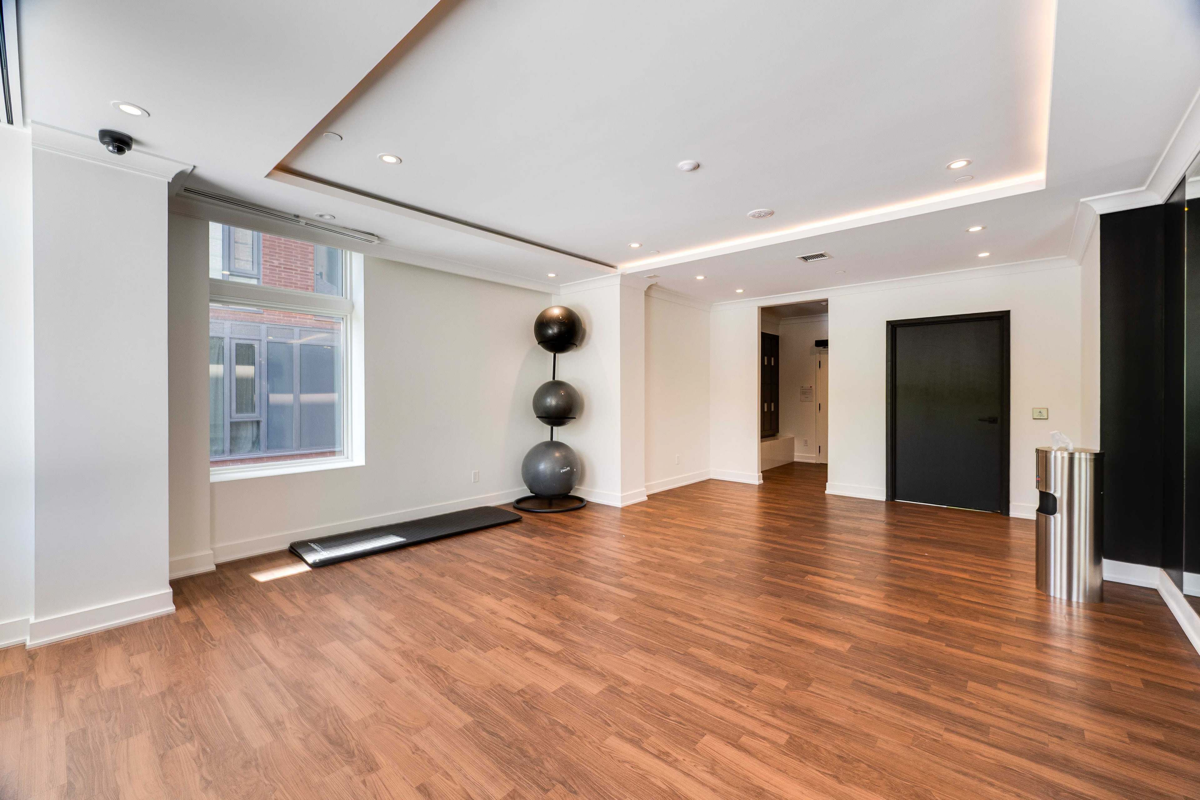 The image shows a spacious room with hardwood flooring, featuring a large window, a black exercise mat, two exercise balls stacked in a corner, and a metallic trash can.