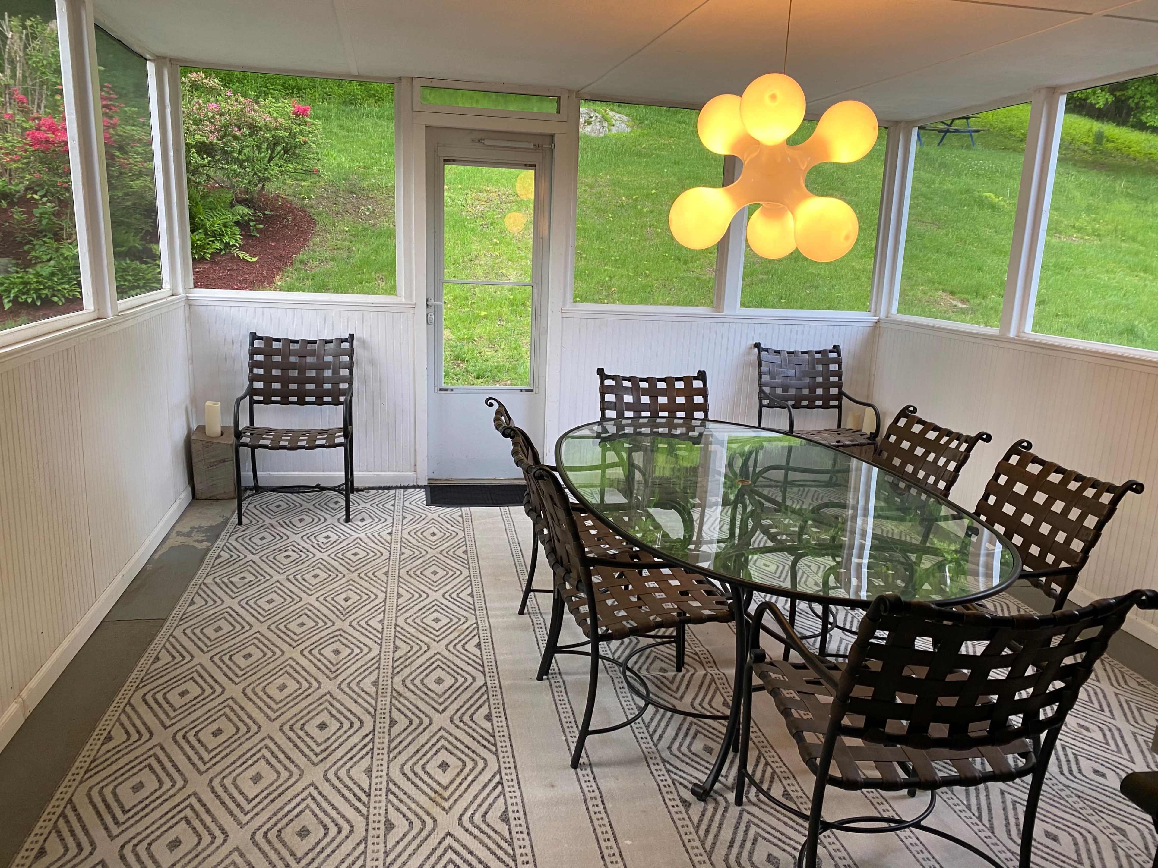 A sunroom featuring a glass dining table surrounded by six black metal chairs and a large light fixture above.