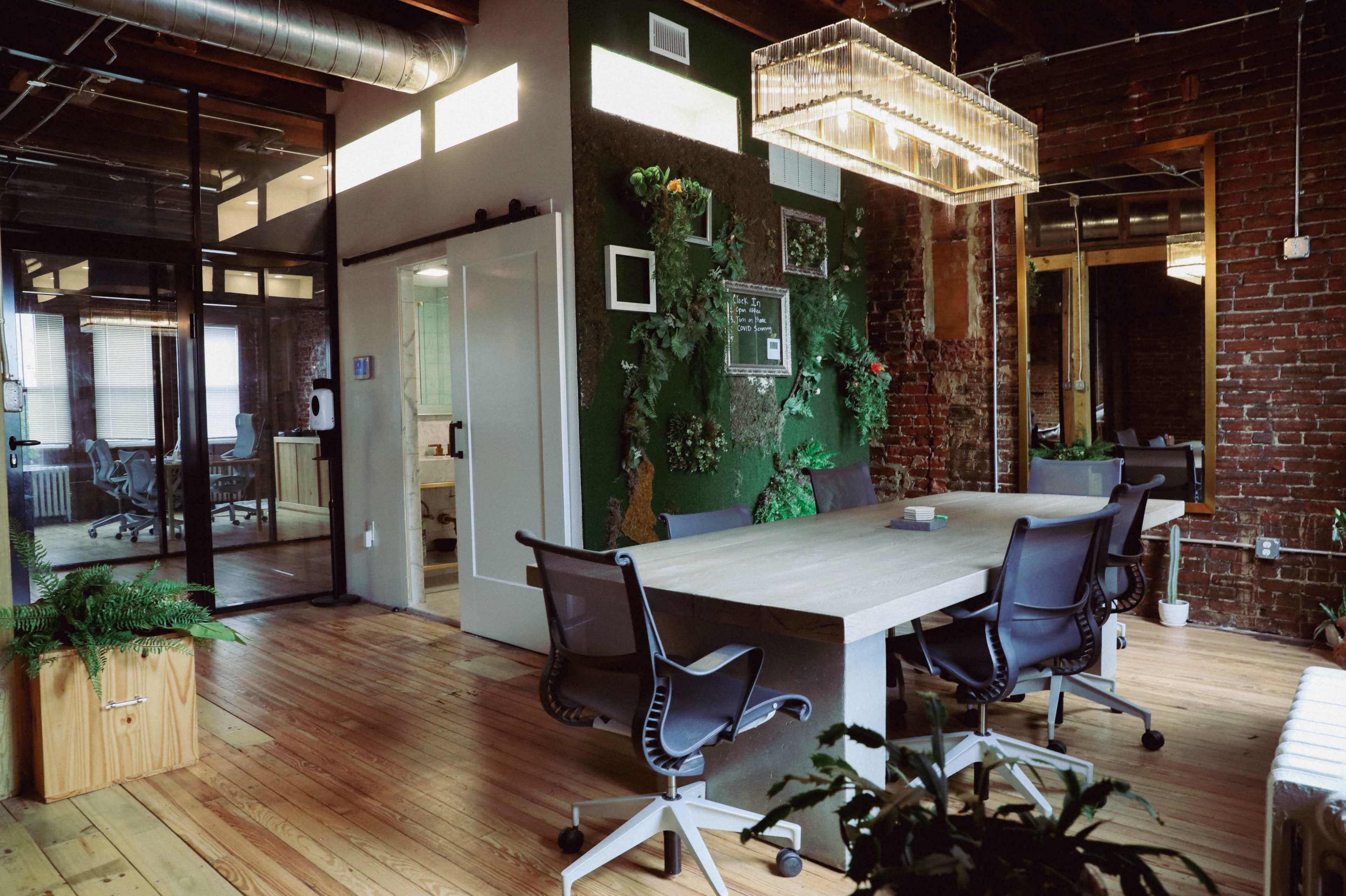 A modern office space featuring a large wooden table surrounded by black chairs, with exposed brick walls and greenery decor.
