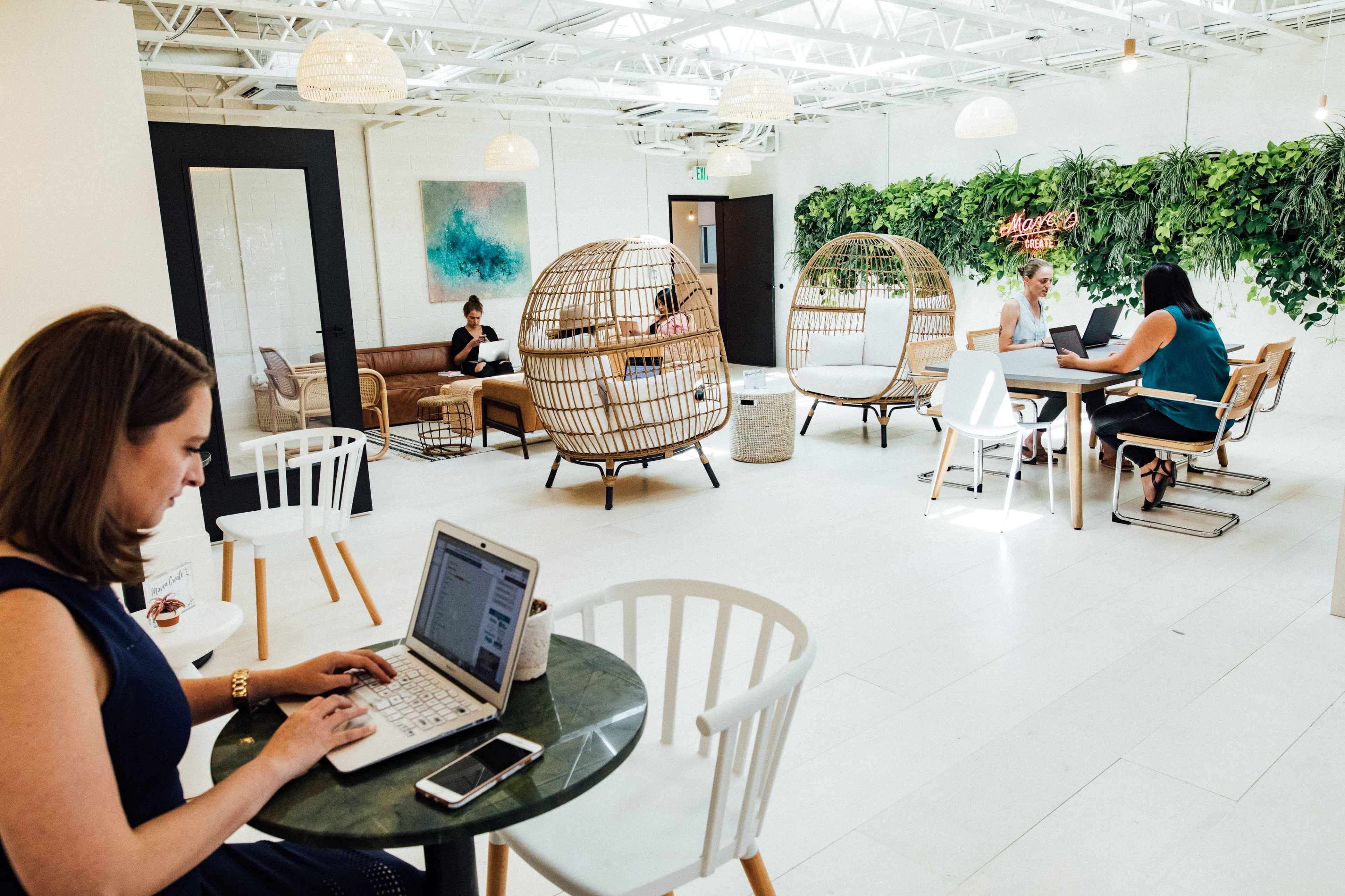 A bright co-working space features various seating arrangements, including wicker chairs, a sleek table, and individuals working on laptops.