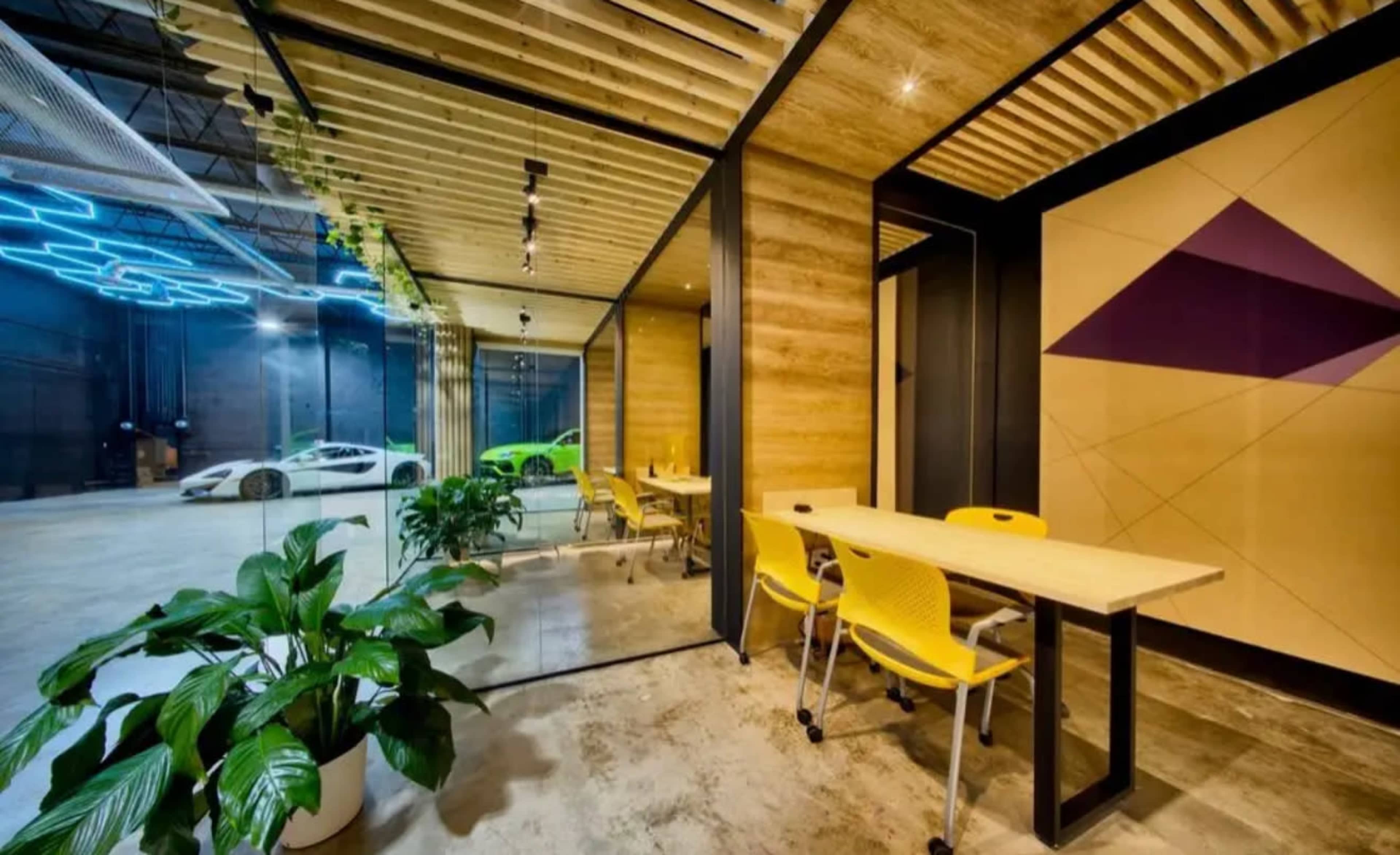 The image shows a modern office space featuring a glass-walled meeting room, a table with yellow chairs, and luxury sports cars visible in the background.