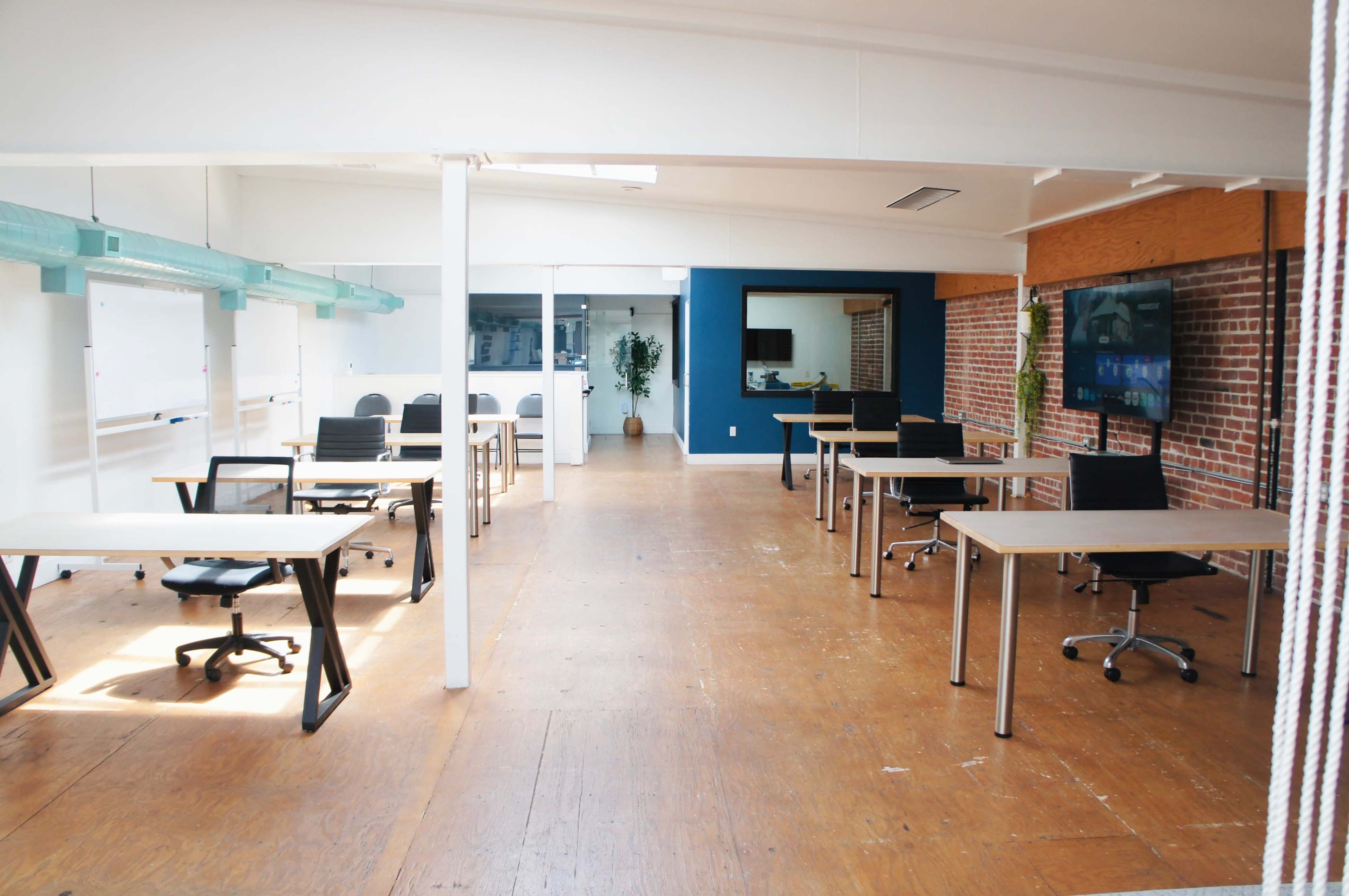 The image shows a modern office space with several desks and chairs arranged in a spacious, well-lit room that has exposed brick walls.