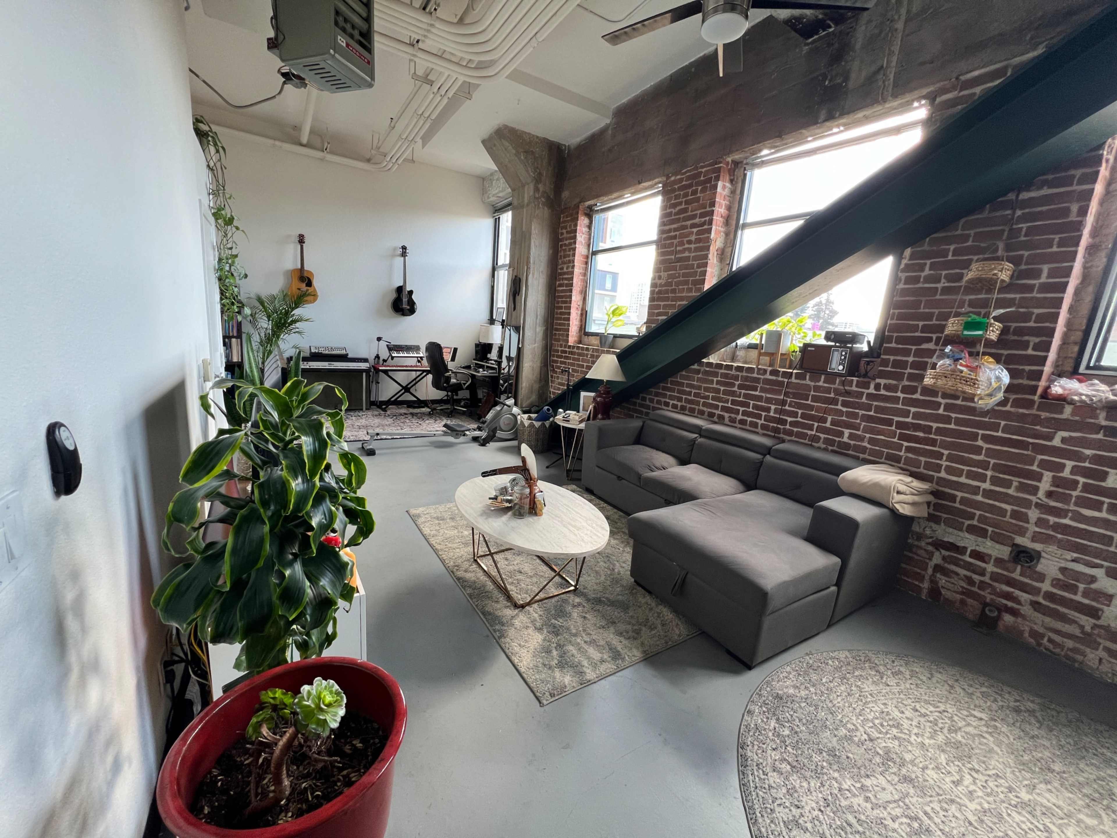 The image shows a loft-style living space with exposed brick walls, a sectional sofa, a coffee table, and a workspace area featuring a desk and guitars.