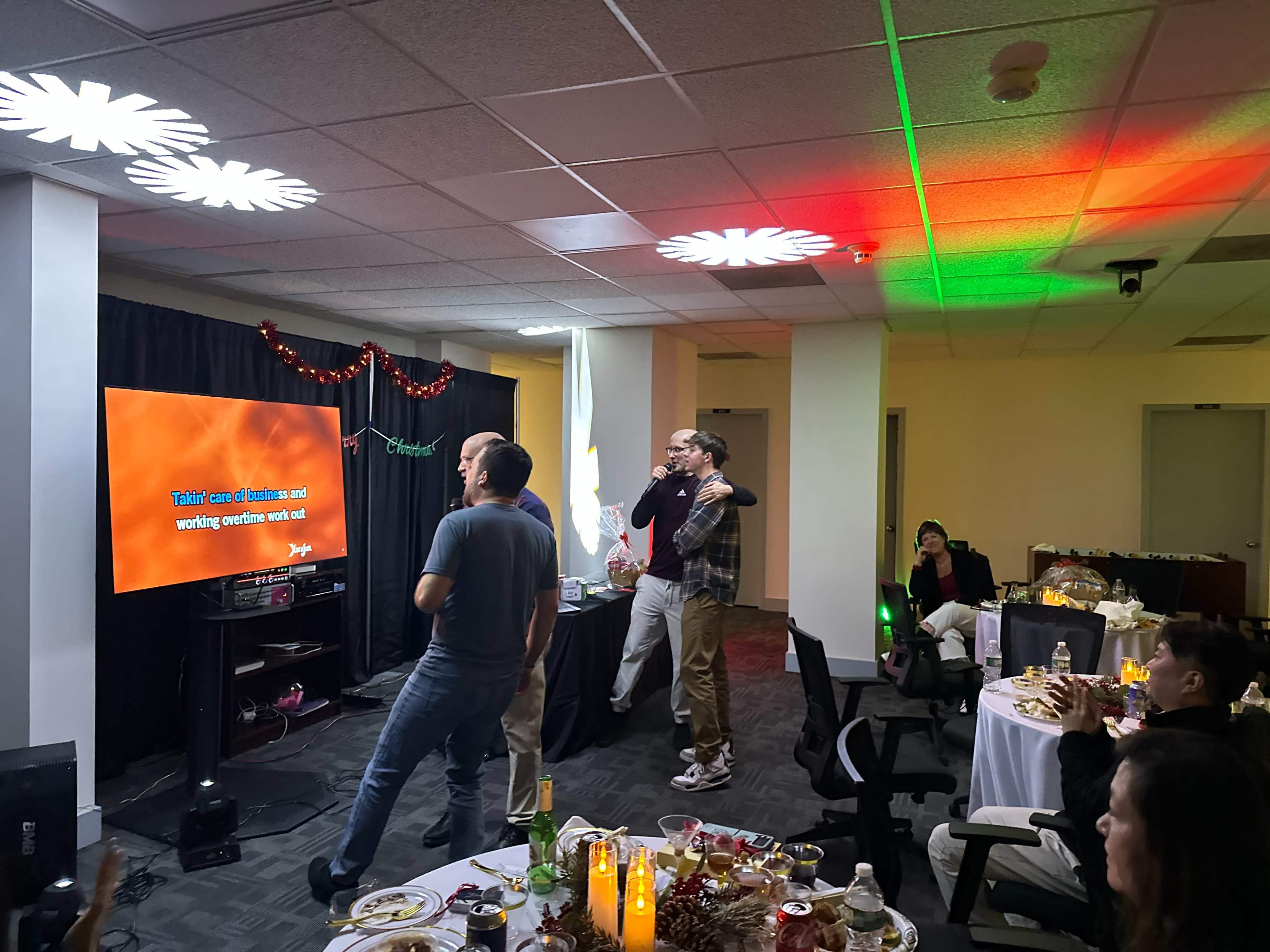 A group of people are singing karaoke in a decorated office space with a large screen displaying lyrics and a festive atmosphere.