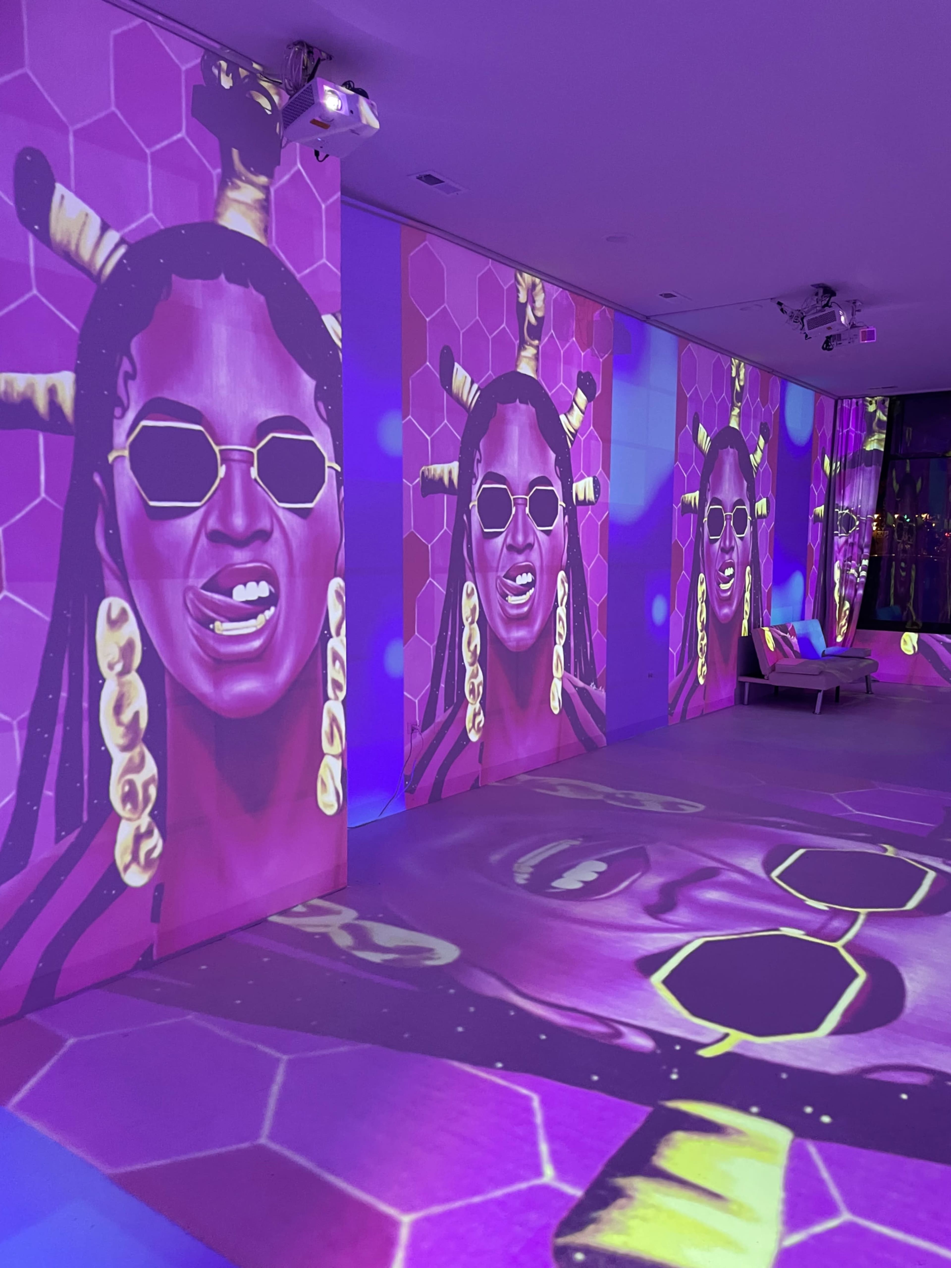 A vibrant, art-filled room with large, bold portraits of a woman projected onto the walls, accompanied by colorful lighting effects.