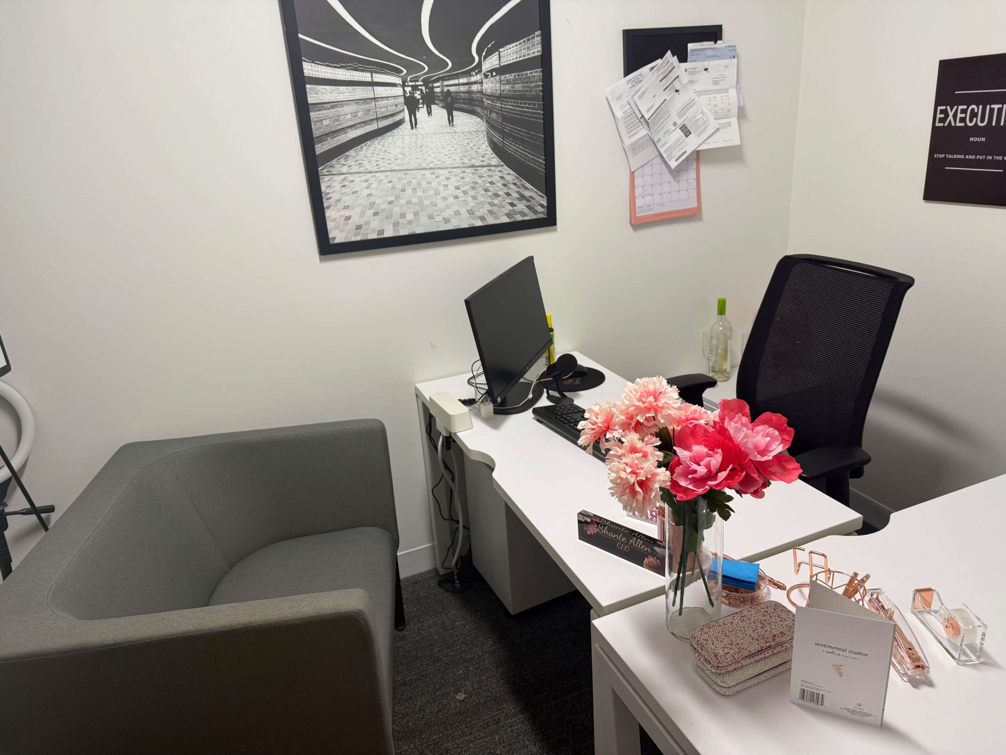 The image shows a small office space with a desk, a computer, a chair, a plant, and a flower arrangement on a table, along with various papers and a framed black-and-white photo on the wall.