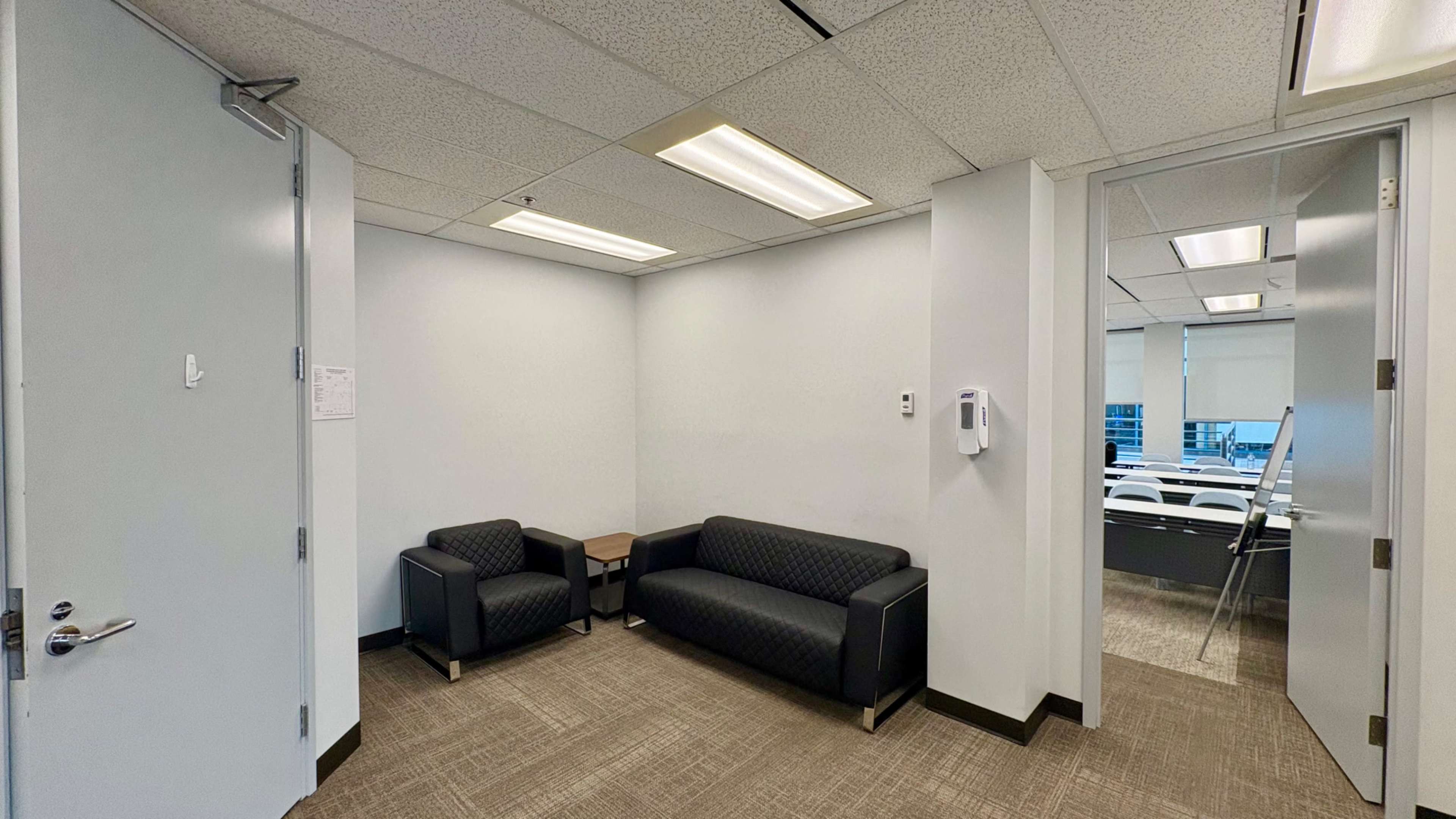 The image shows a small waiting area with two black sofas and a side table, adjacent to an office space with a door leading to a conference room.
