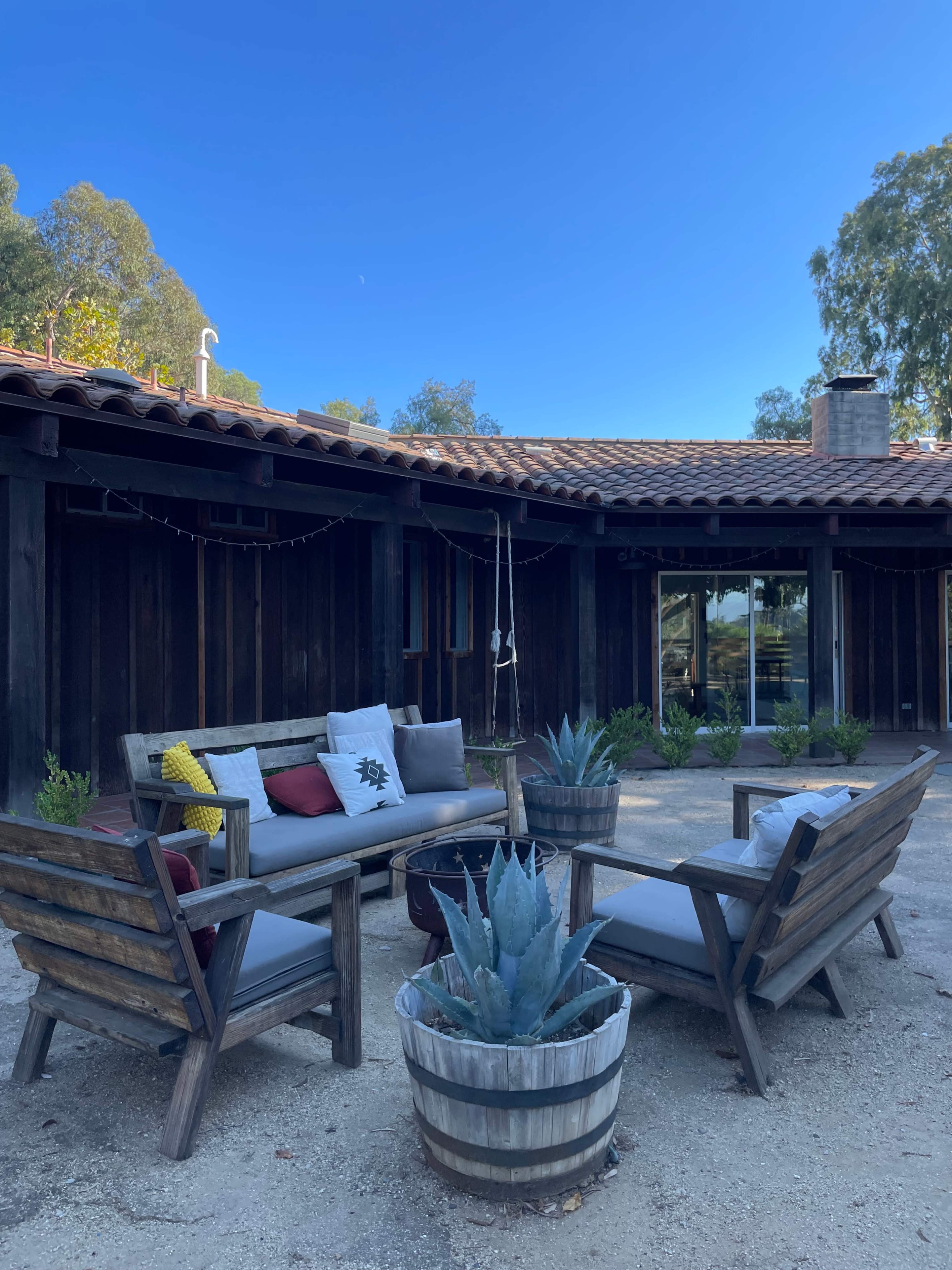 A cozy outdoor seating area features a sofa and two chairs arranged around a central planter filled with an agave plant, set in front of a rustic wooden building.
