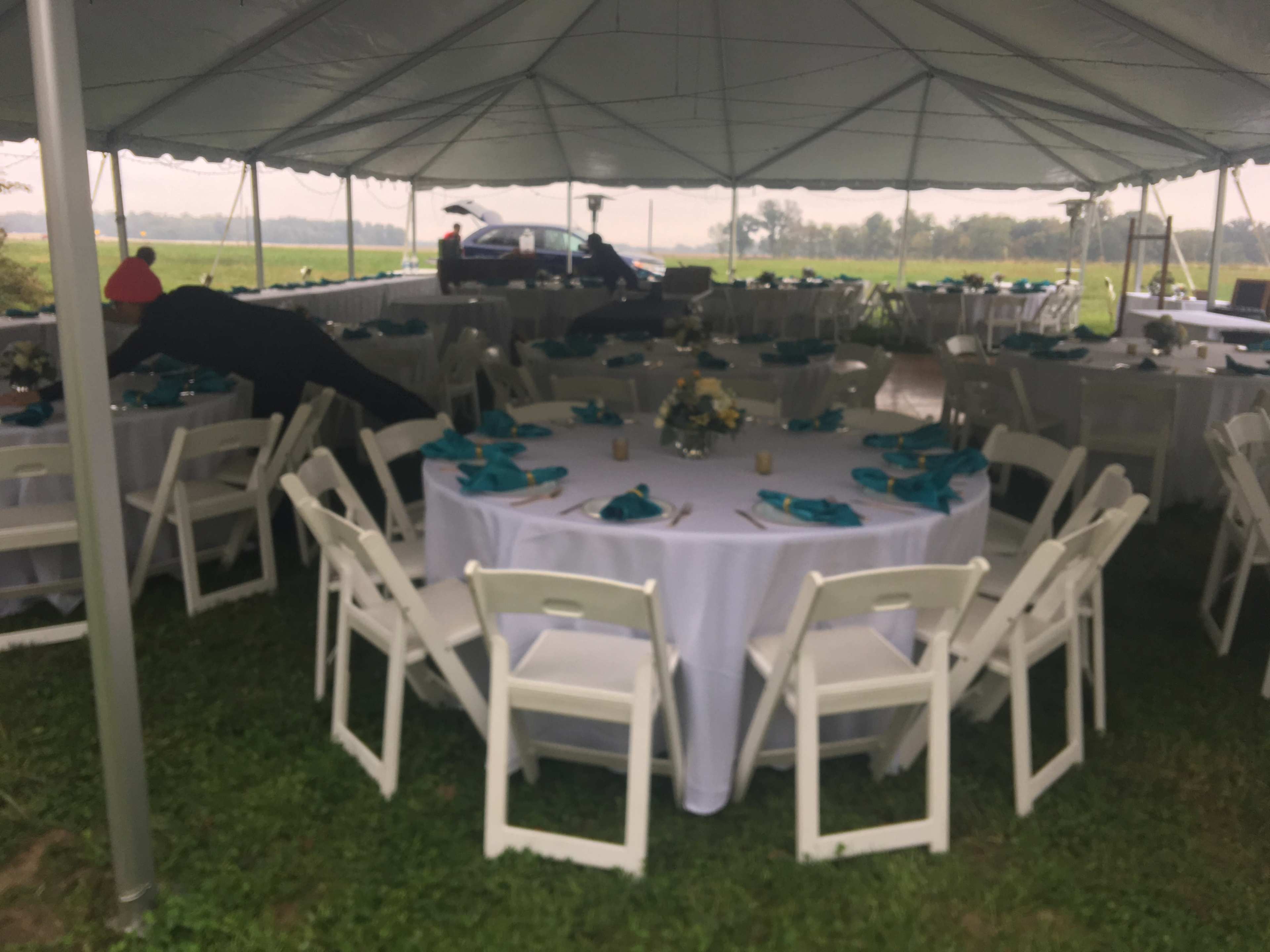 A spacious tent with neatly arranged round tables covered in white tablecloths and teal napkins, surrounded by chairs, set up for an outdoor event.