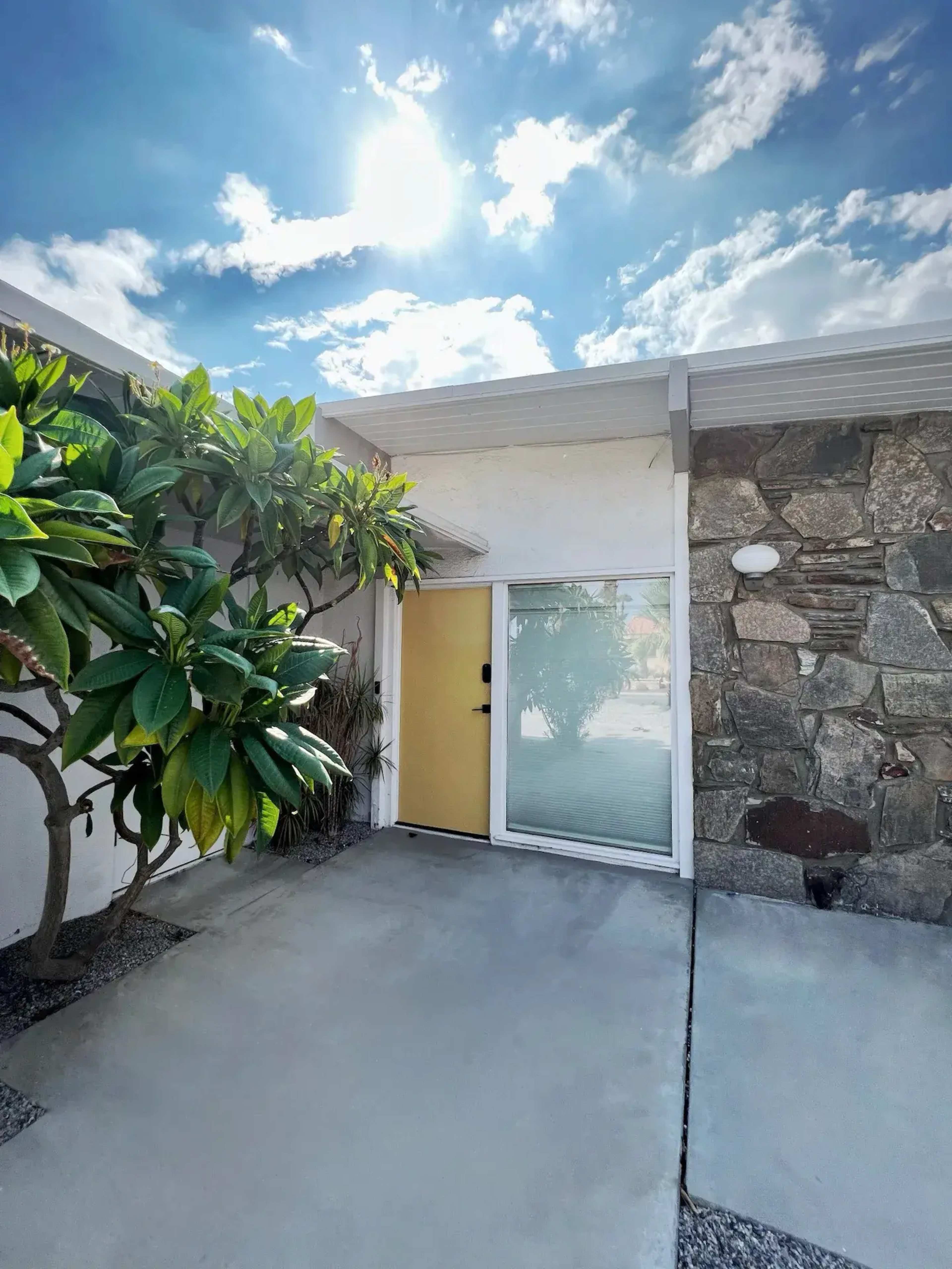 A modern building entrance features a glass door framed by a stone wall and a sunlit sky with scattered clouds.
