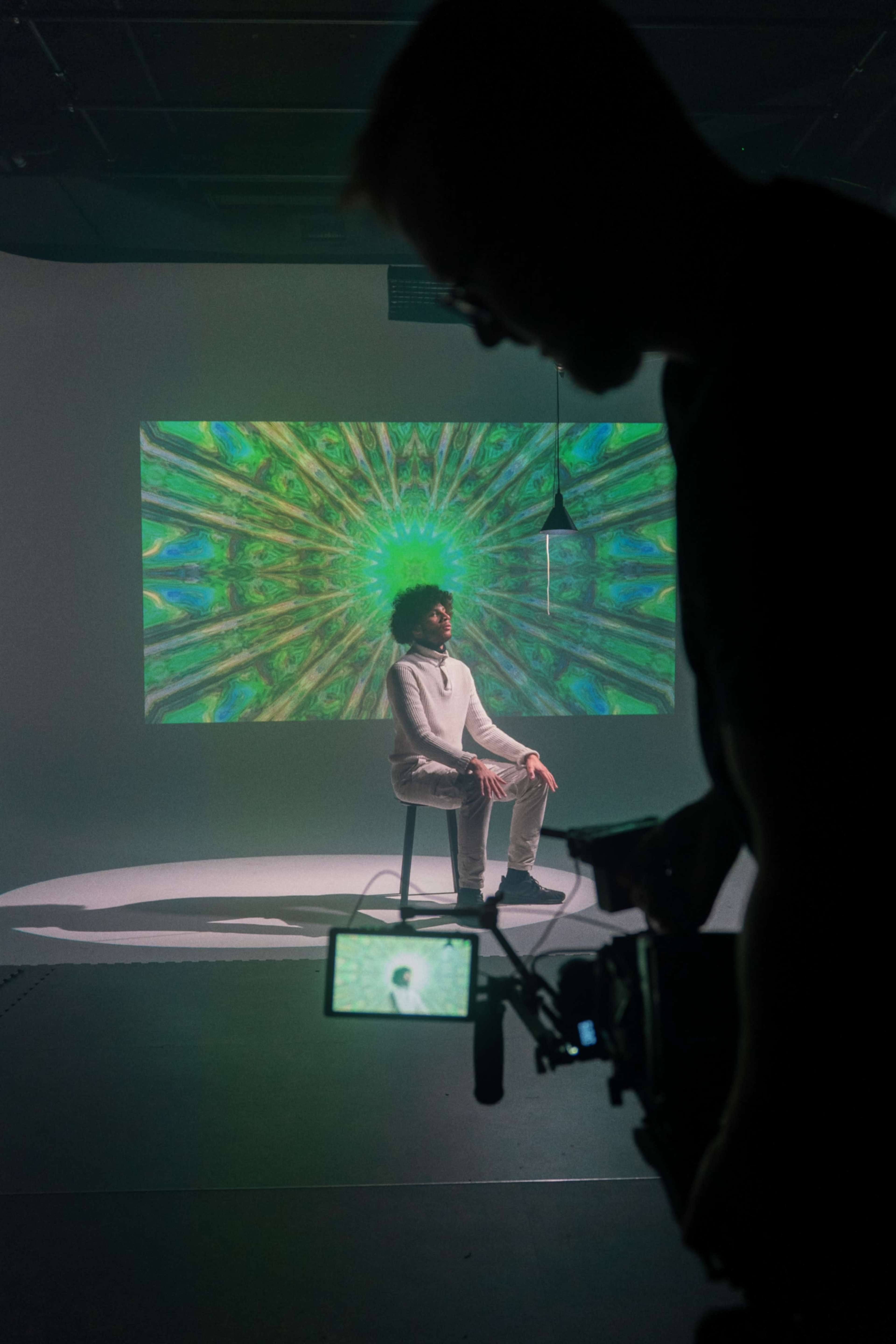 A person sits on a stool in front of a colorful, abstract backdrop while another individual, silhouetted in the foreground, holds a camera.
