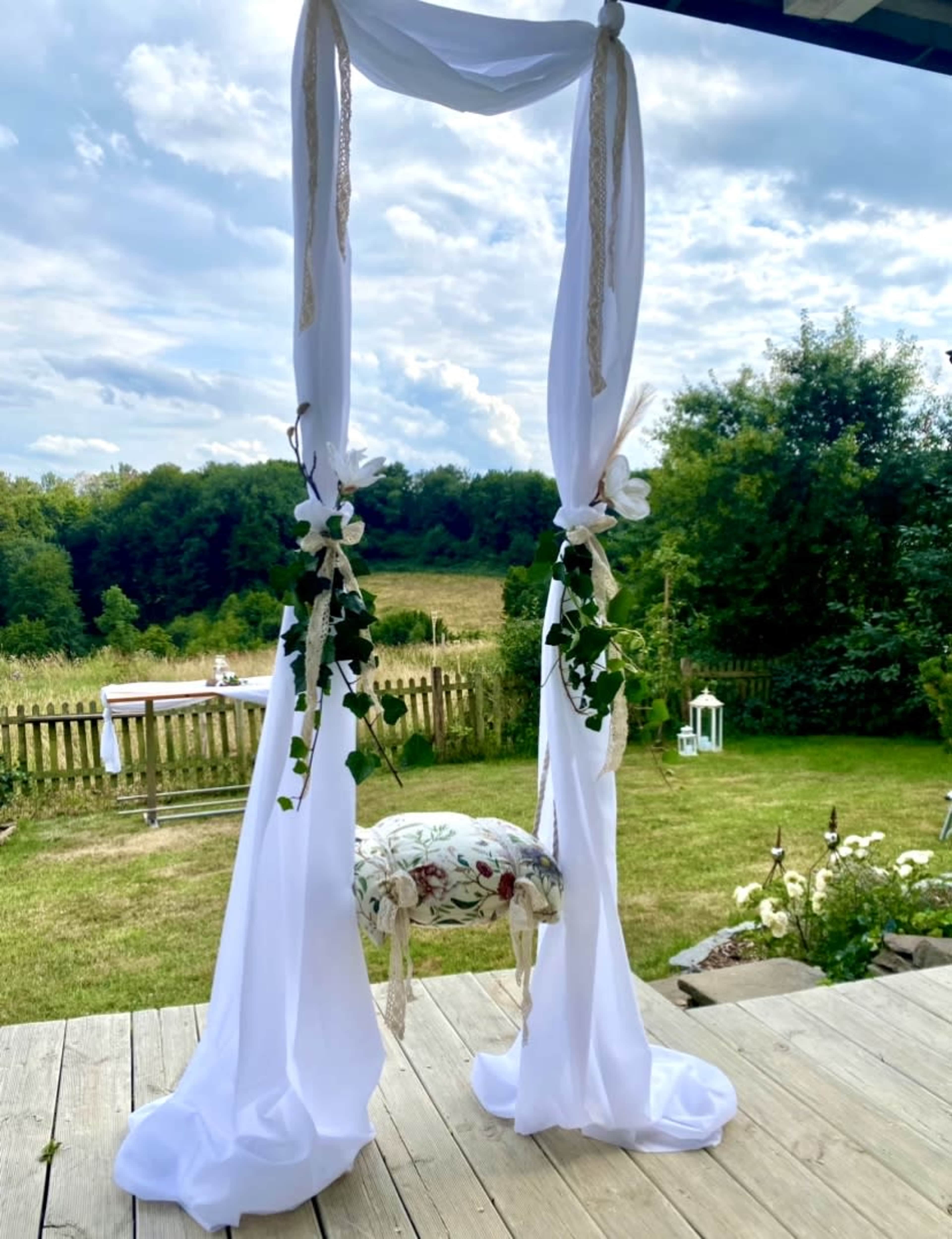 A white draped arch with floral accents stands in a grassy area, surrounded by trees under a cloudy sky.