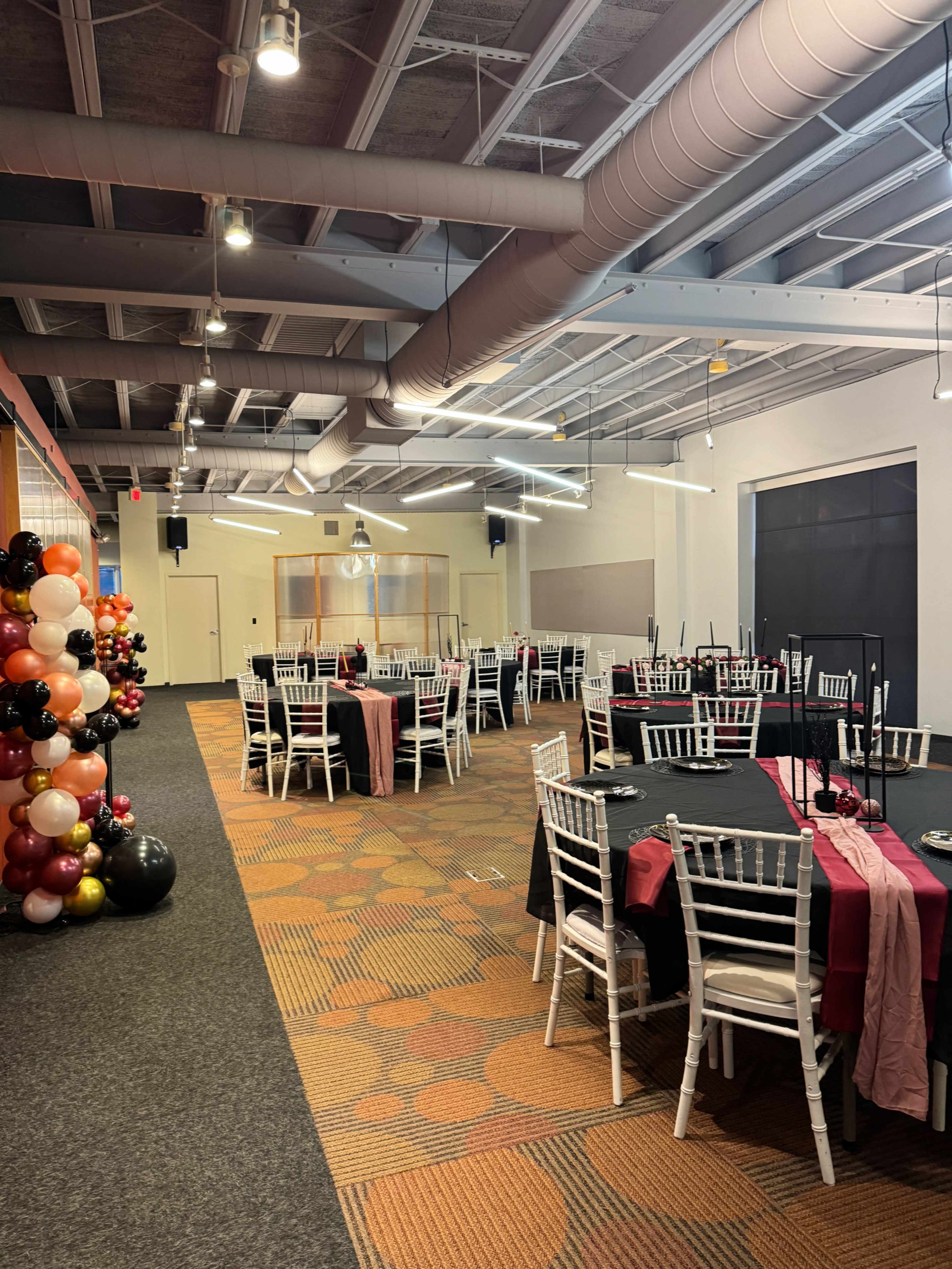 A spacious event room with neatly arranged tables covered in black and burgundy tablecloths, surrounded by white chairs, and decorated with a colorful balloon arch in one corner.