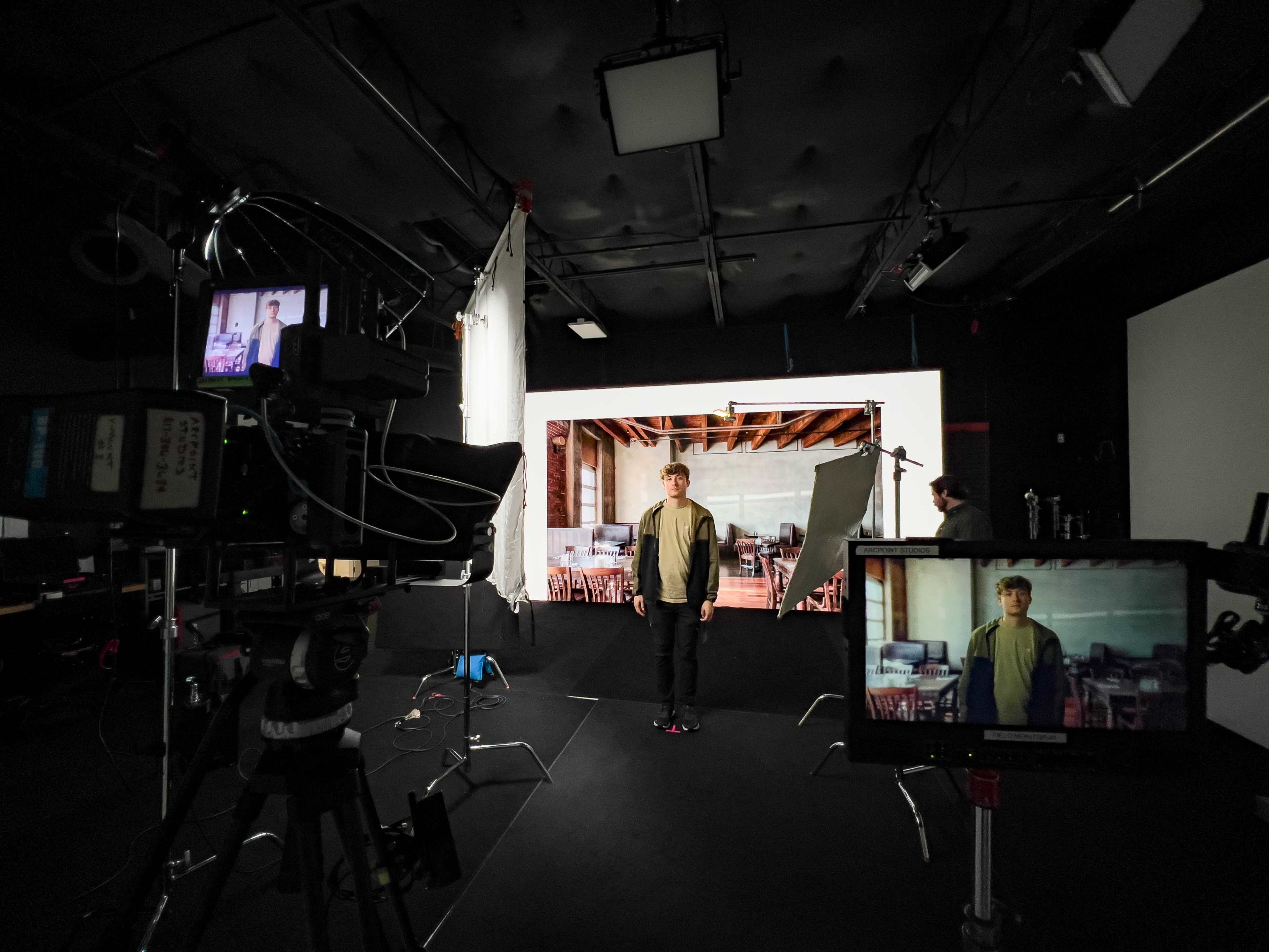 A person stands in a studio surrounded by cameras and lighting equipment, with a large backdrop featuring a room setup behind them.