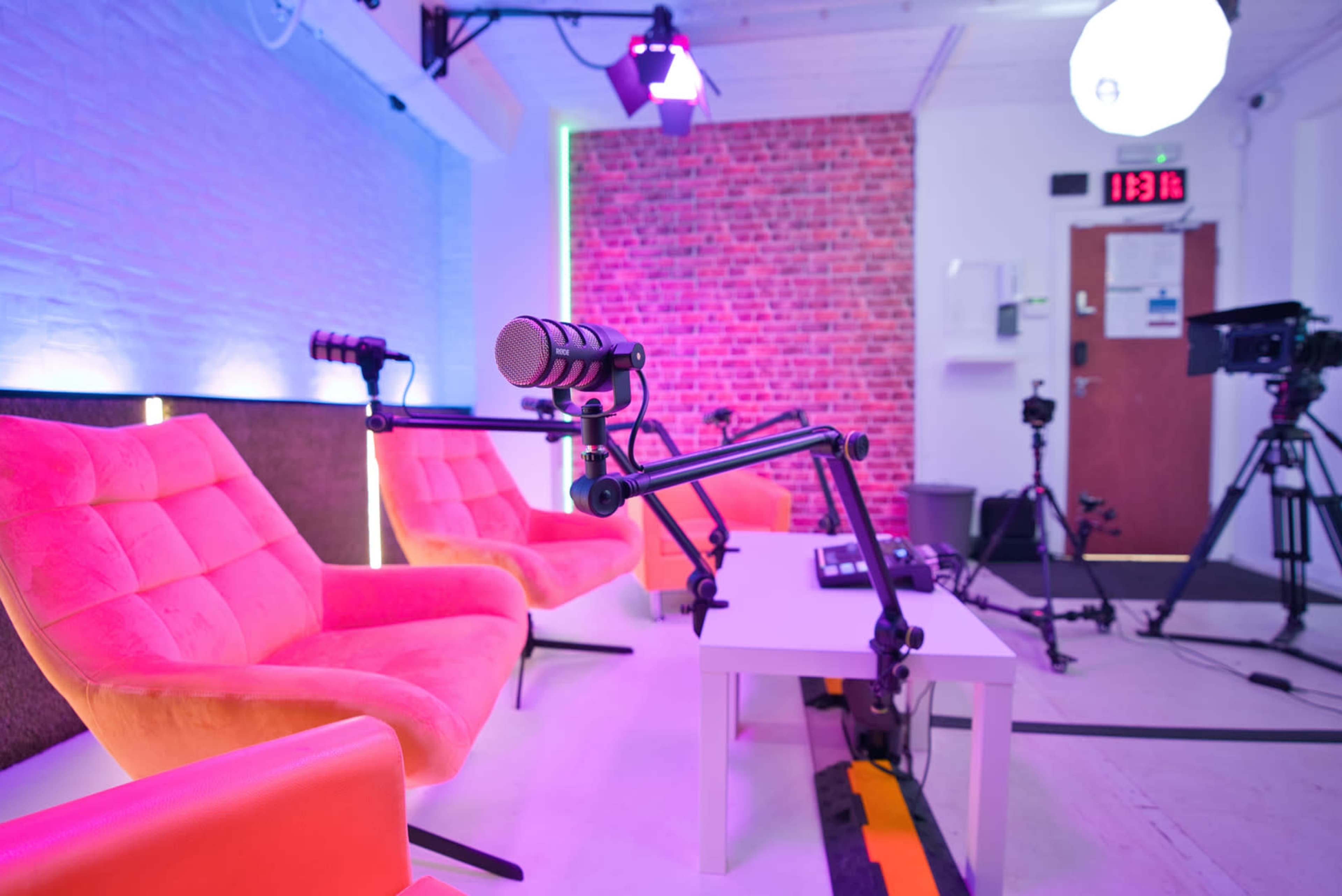 A modern recording studio features two pink chairs, microphone setups, and various filming equipment against a brick wall backdrop.