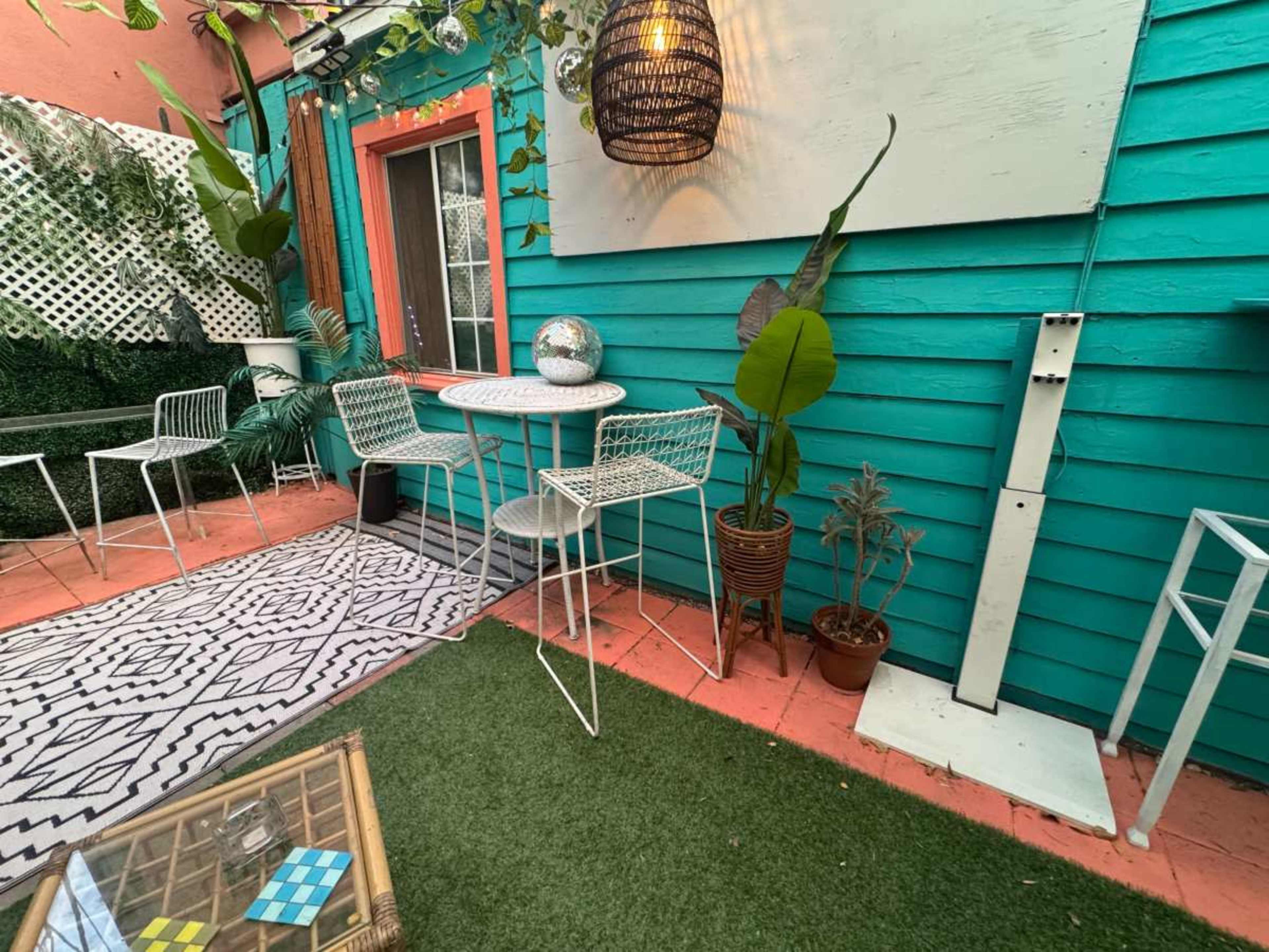 The image shows a colorful outdoor space featuring a small round table with four white chairs, surrounded by green plants and decorative elements against a turquoise wall.