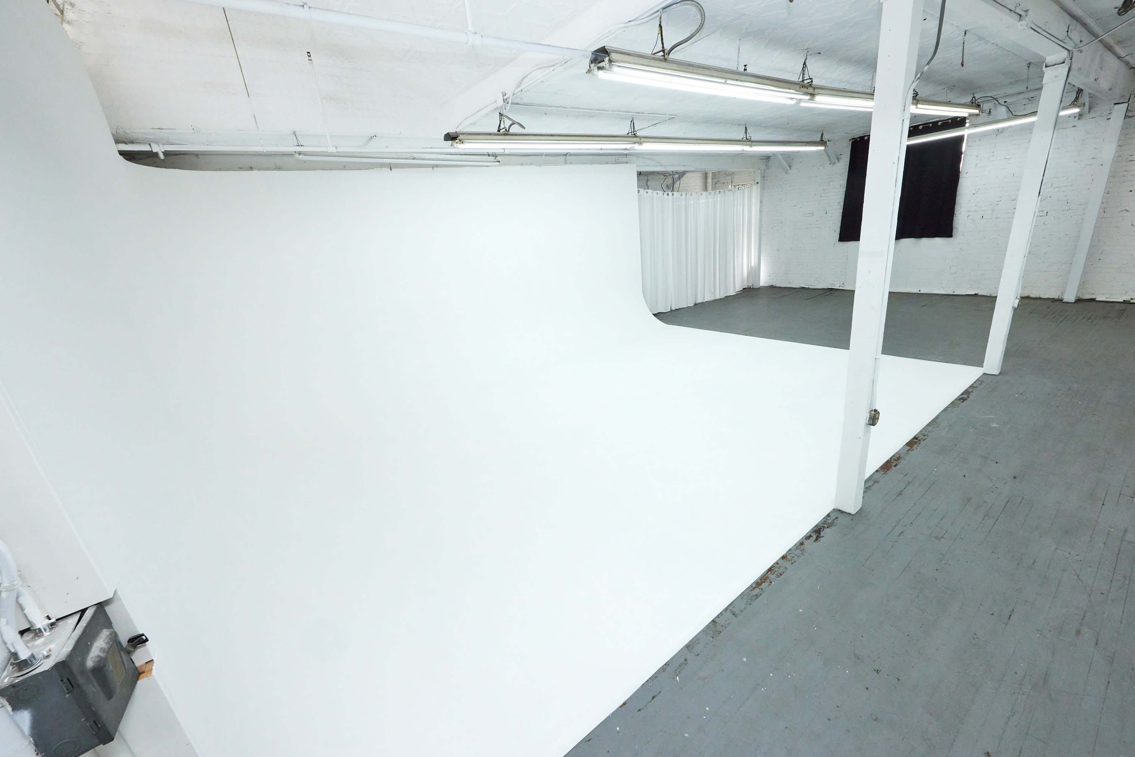 The image shows a spacious photography studio with a seamless white backdrop and a gray wooden floor.
