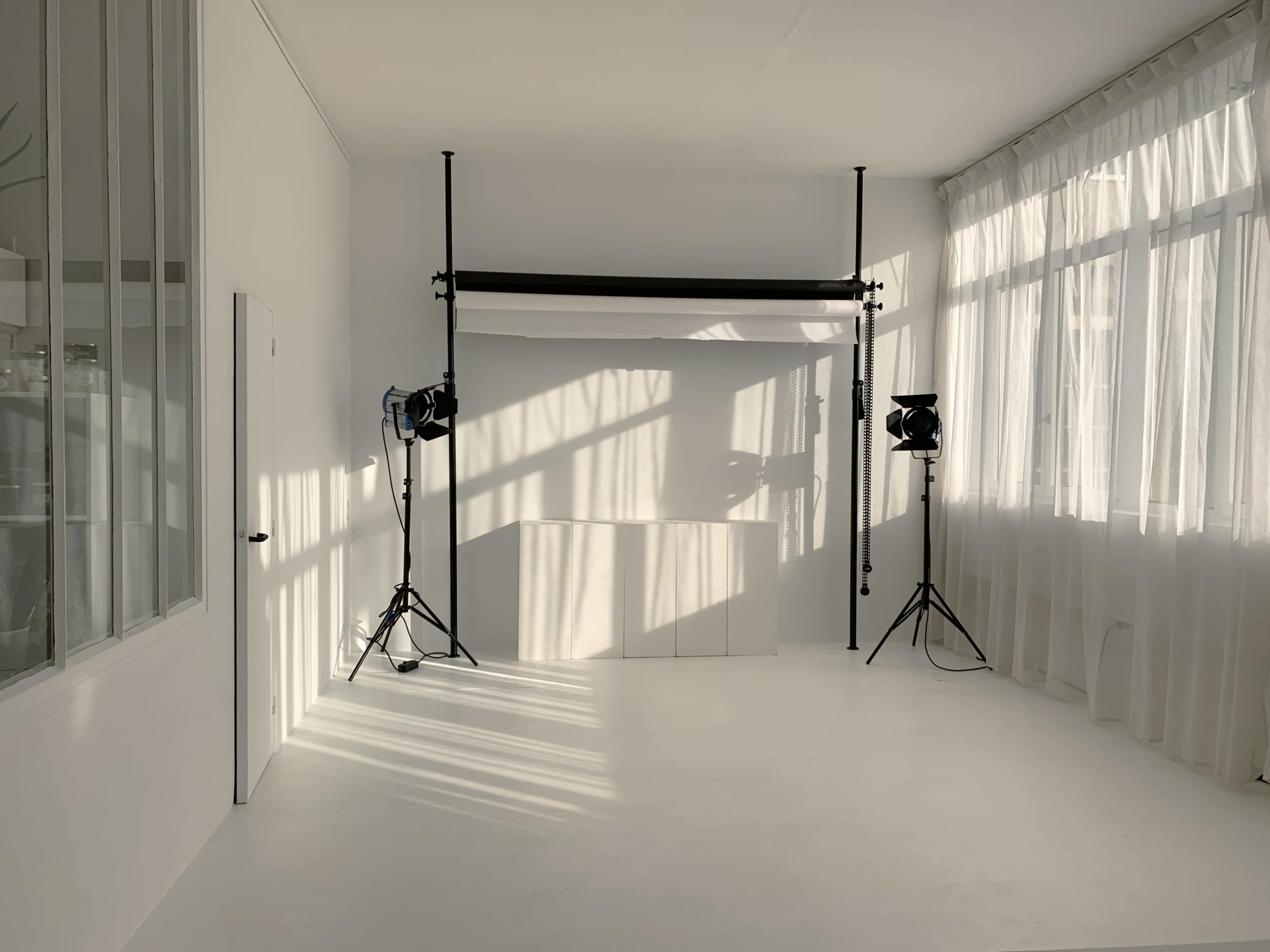 A bright photography studio with white walls, a backdrop stand, and two light stands casting shadows on the floor.