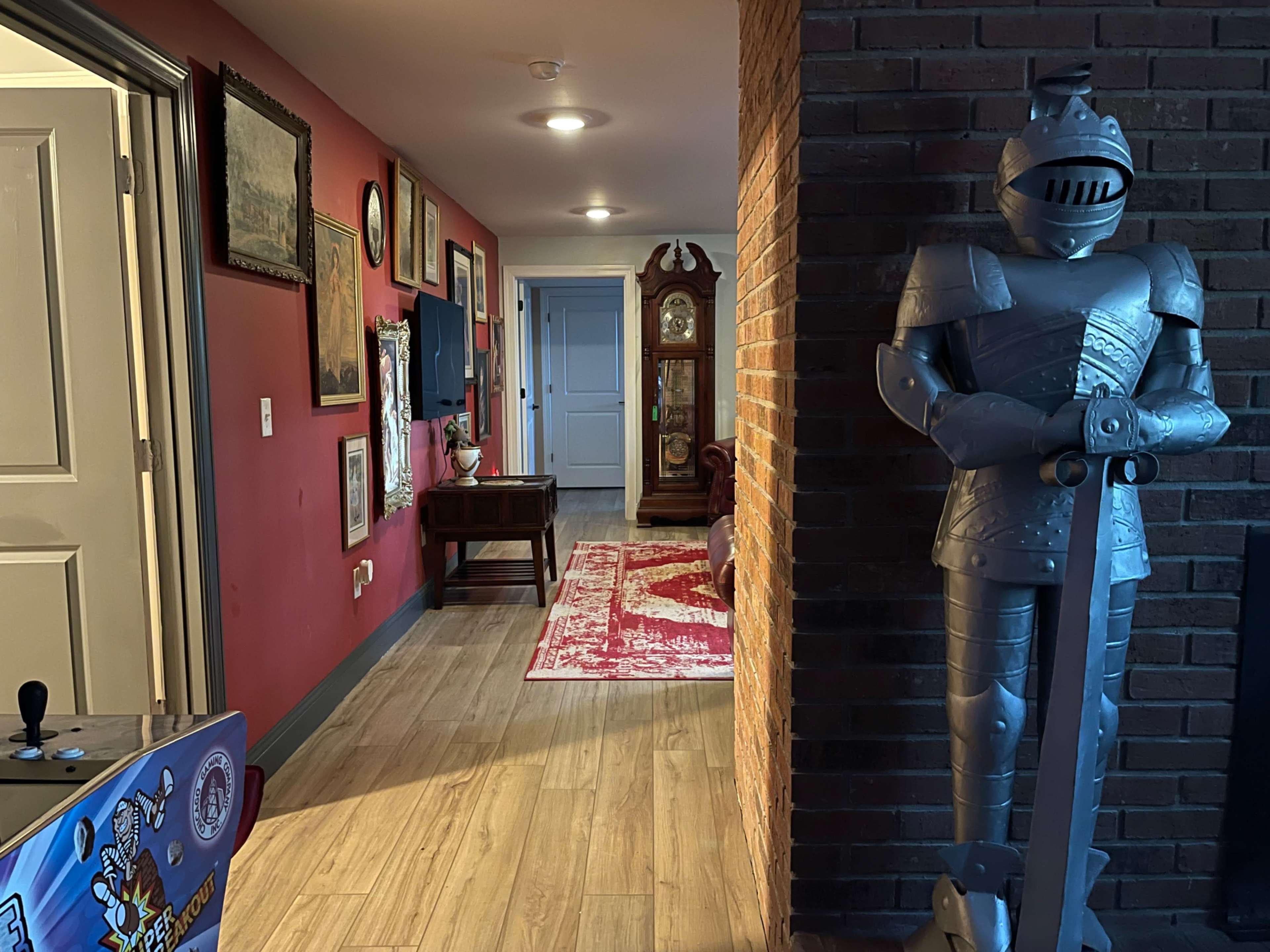 The image shows a hallway adorned with framed artwork, wooden furniture, a grandfather clock, and a suit of armor positioned next to a brick wall.