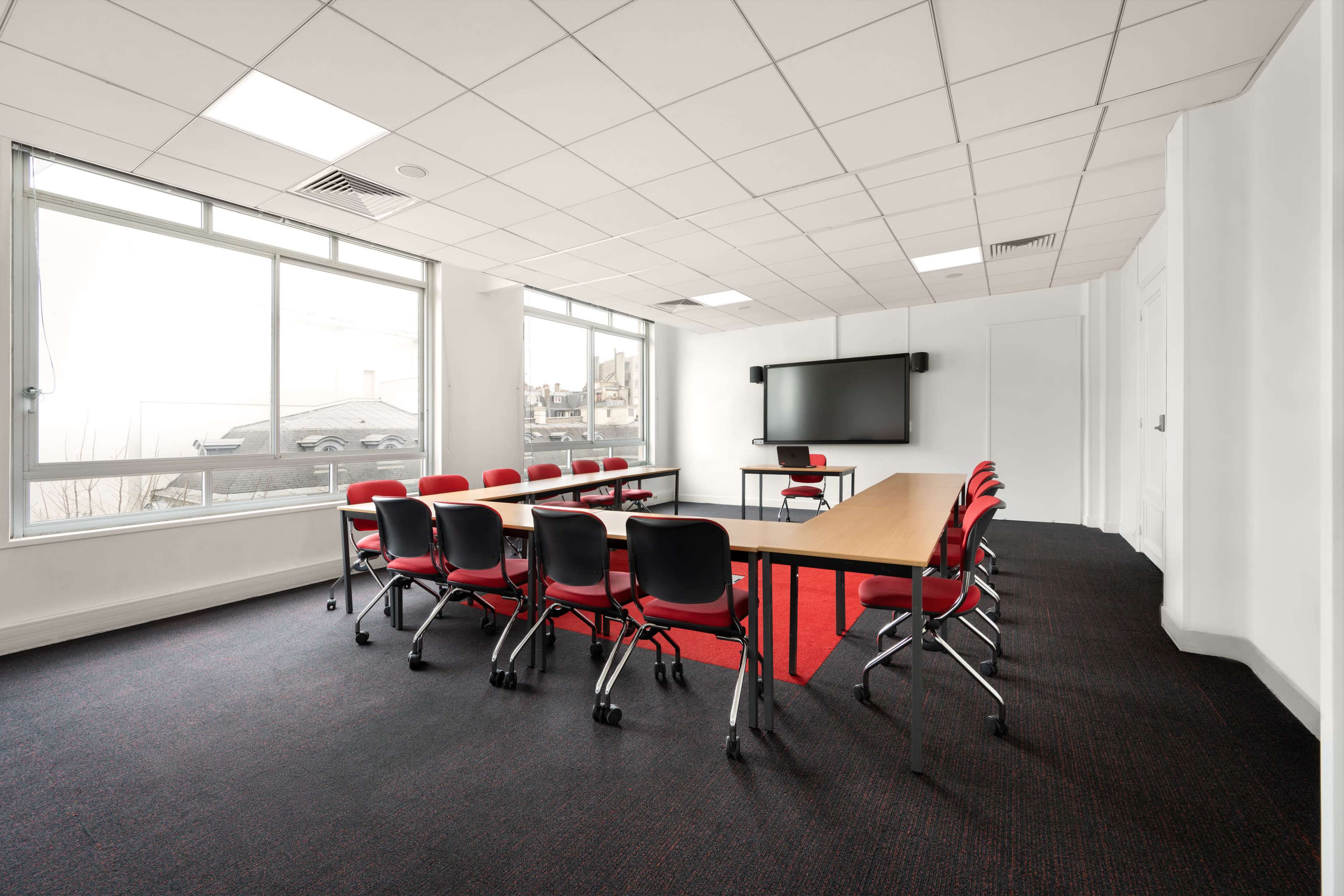 A conference room with a long table surrounded by red chairs, large windows, and a wall-mounted screen.