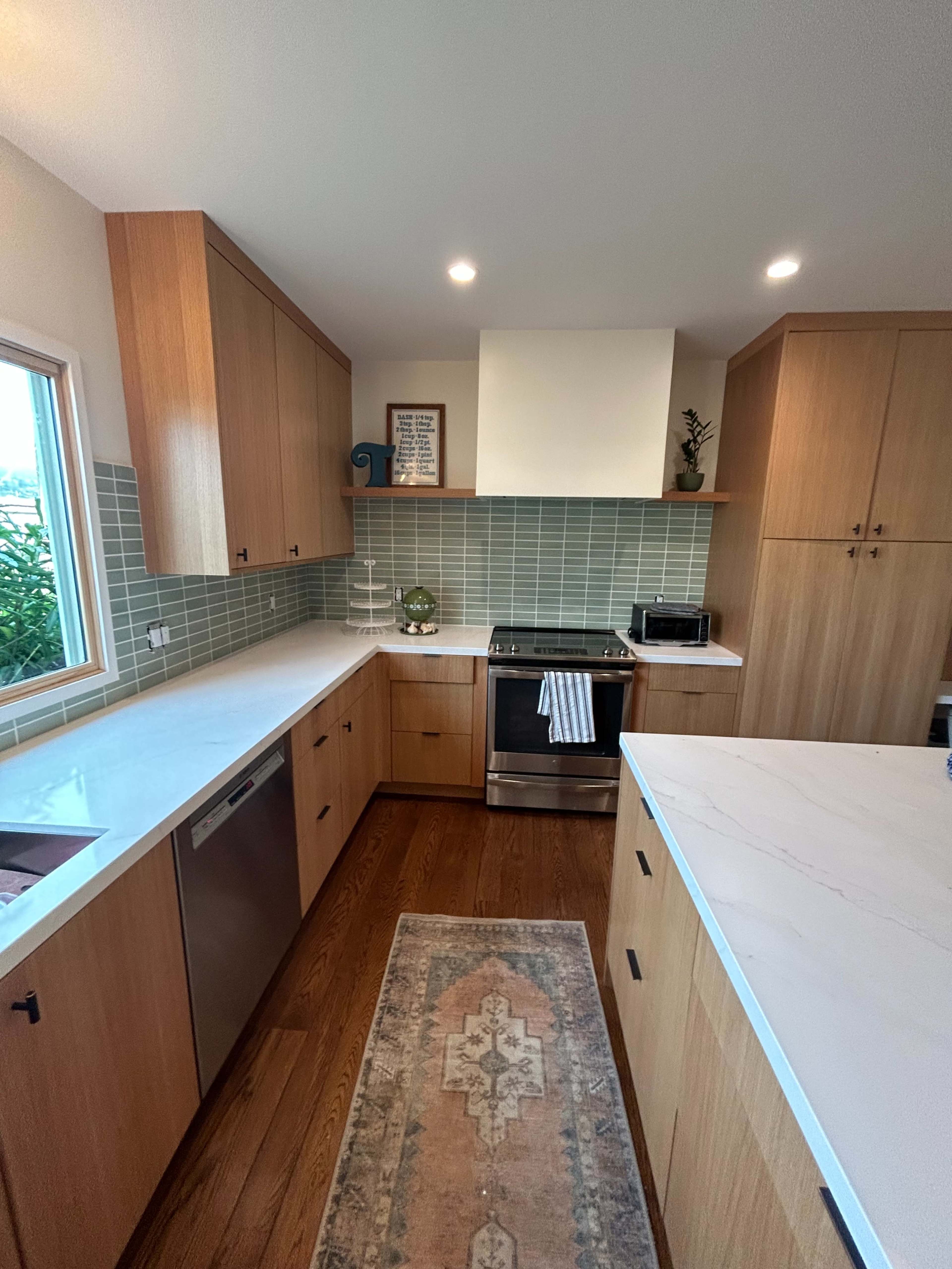 The image shows a modern kitchen featuring wooden cabinetry, a stainless steel oven, and a light-colored countertop with a rug on the wooden floor.