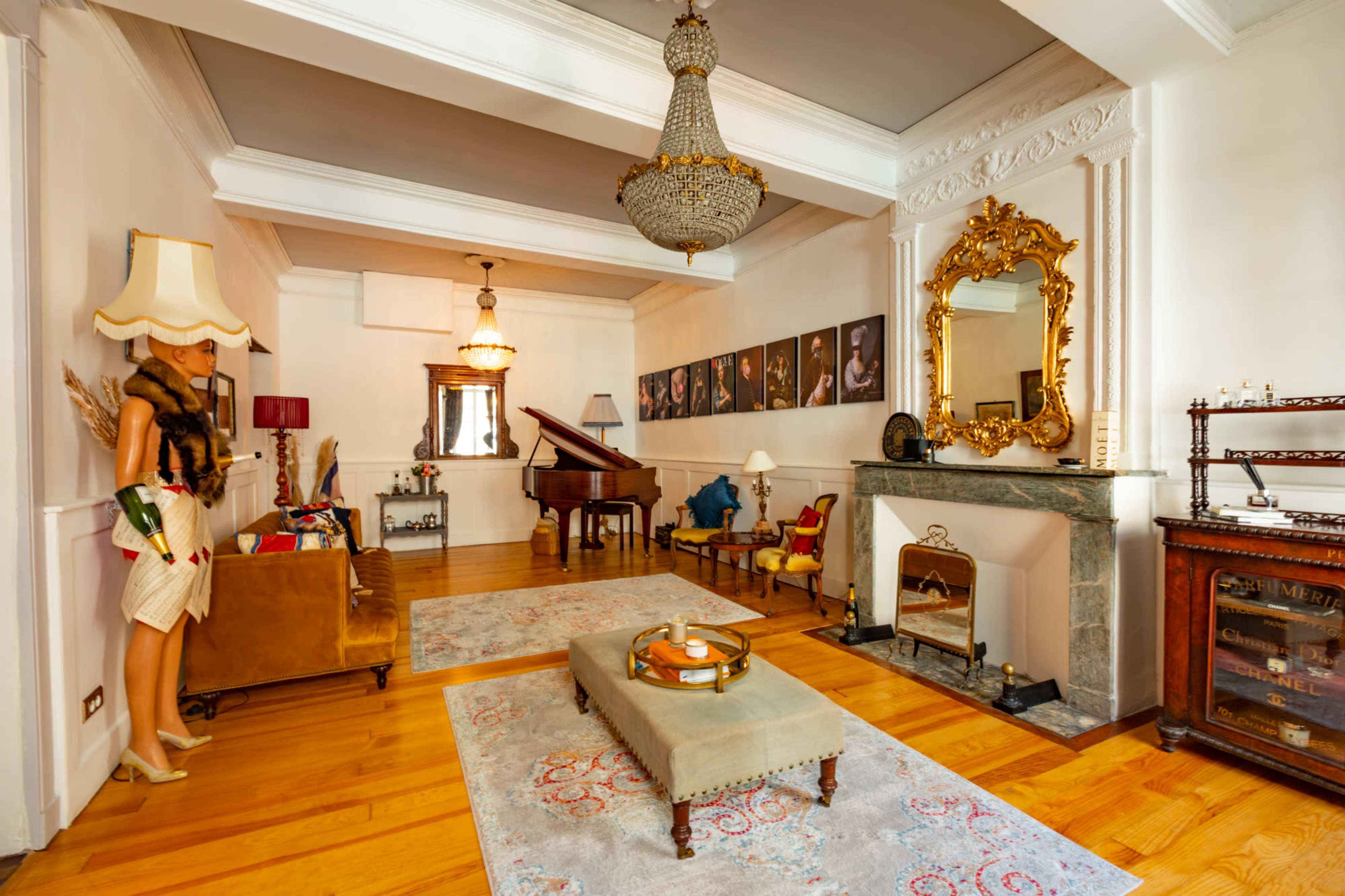 The image shows a spacious, elegantly decorated living room featuring a grand piano, a chandelier, and a mix of vintage furniture and artwork.