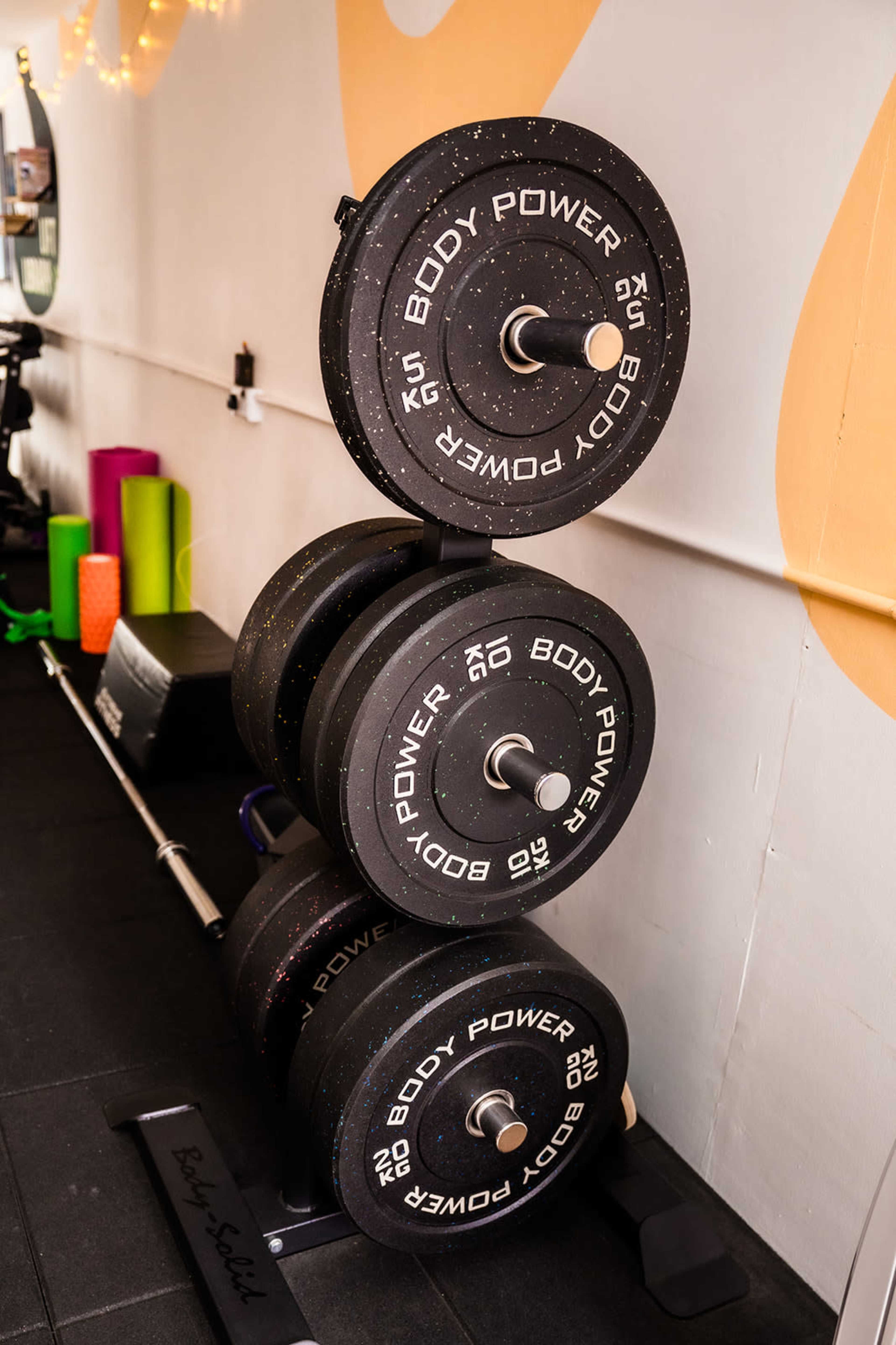 The image shows a three-tier weight rack holding black rubber-coated weight plates labeled "BODY POWER" in a gym setting.