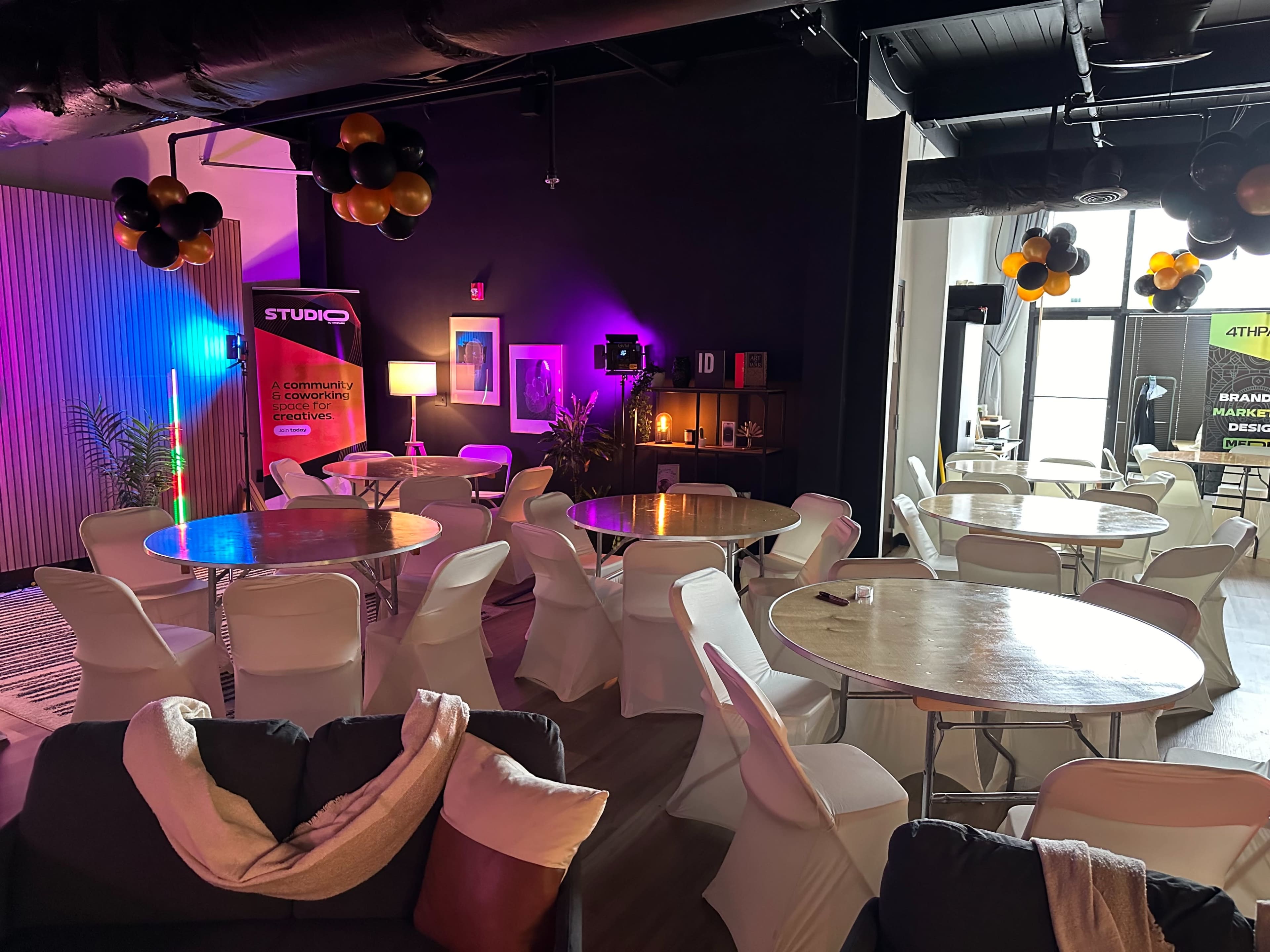 A modern event space with round tables and chairs arranged for a gathering, decorated with balloons and colorful lighting.