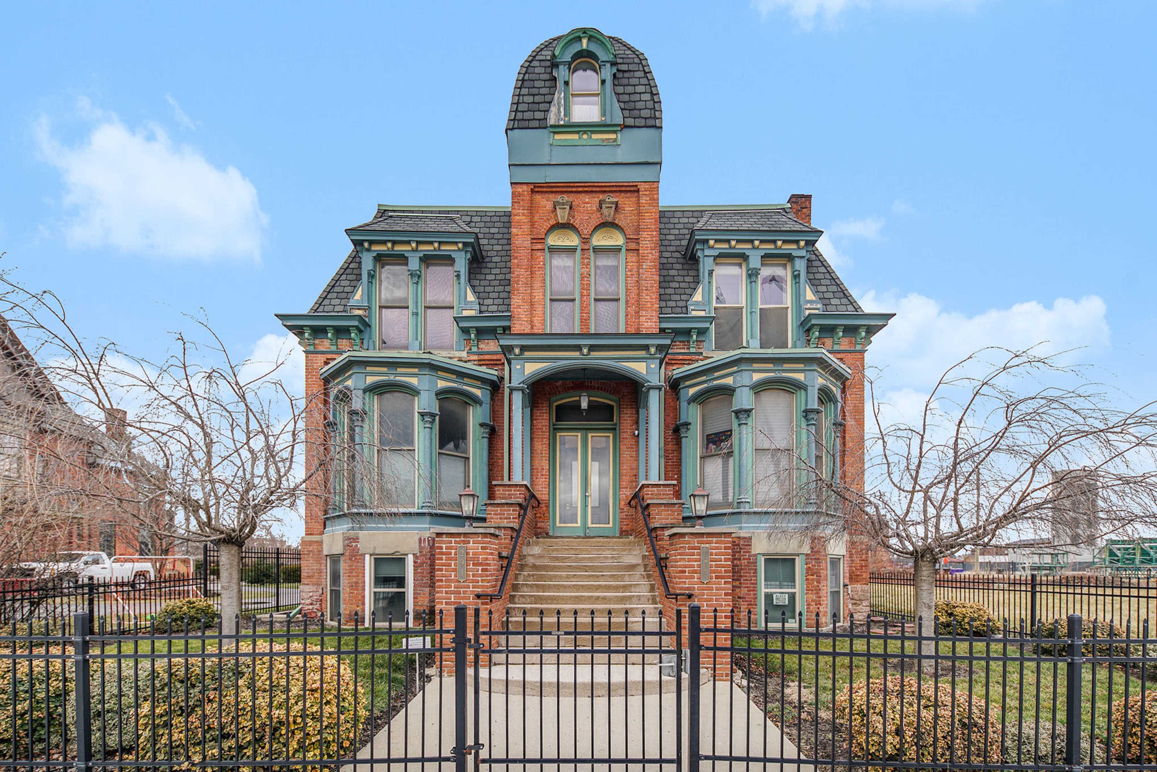A large Victorian-style brick house with a distinctive turret, surrounded by a wrought-iron fence and a landscaped yard.