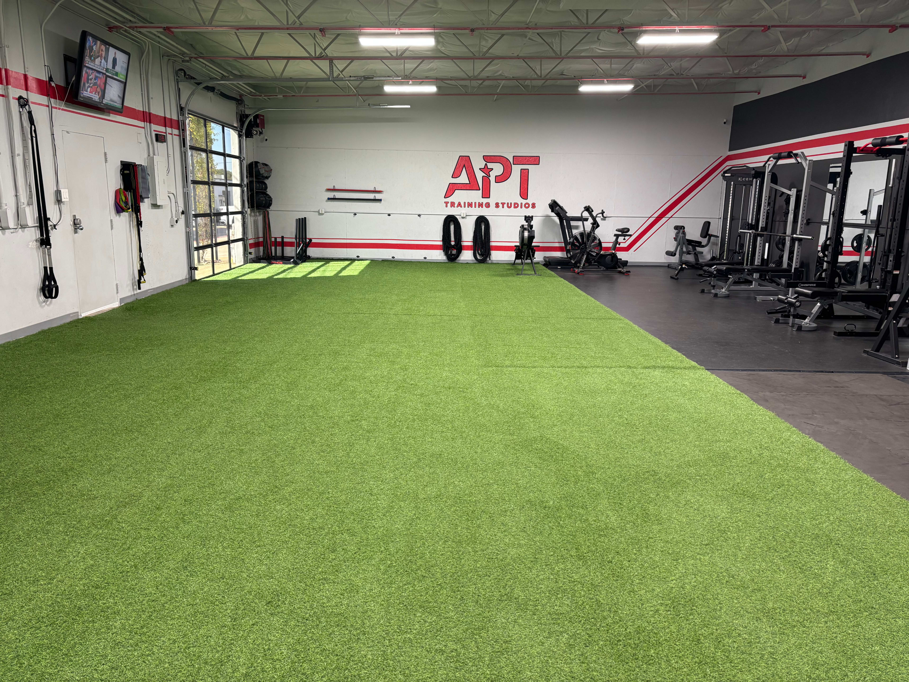 The image shows a training studio featuring a green artificial turf area in the center and various workout equipment along the walls.