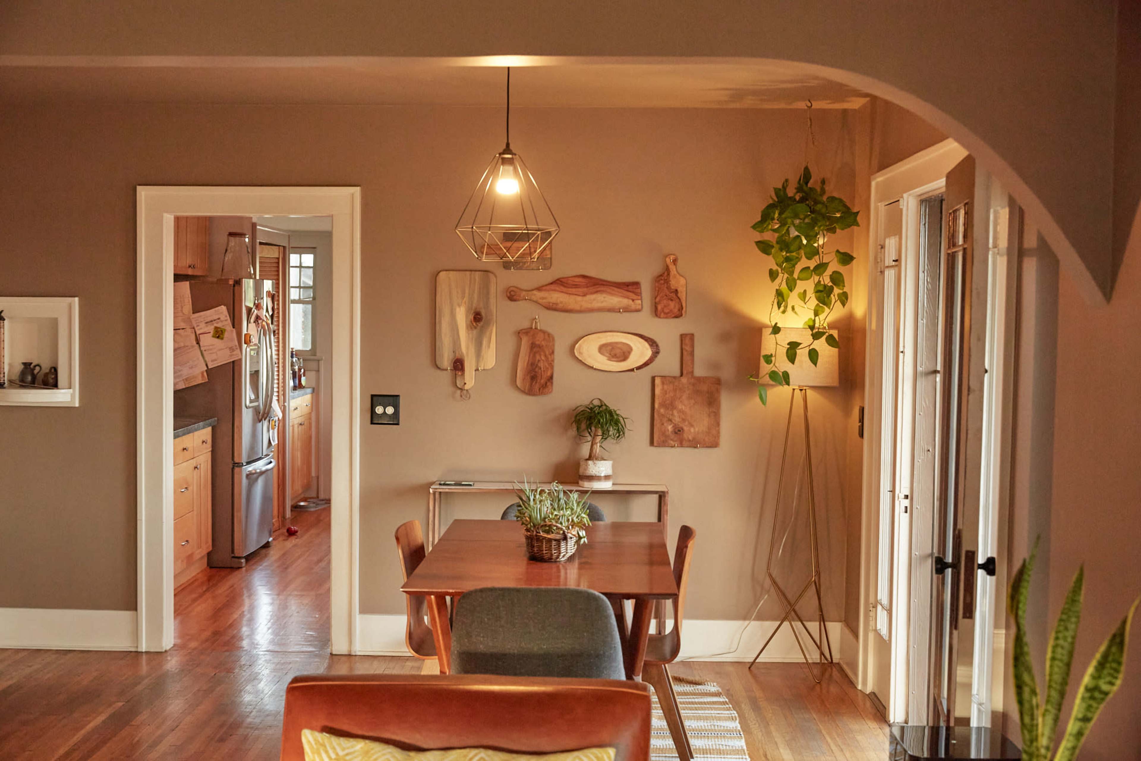 A dining area features a wooden table with chairs, adorned with wall art and a hanging plant, alongside an adjacent kitchen.