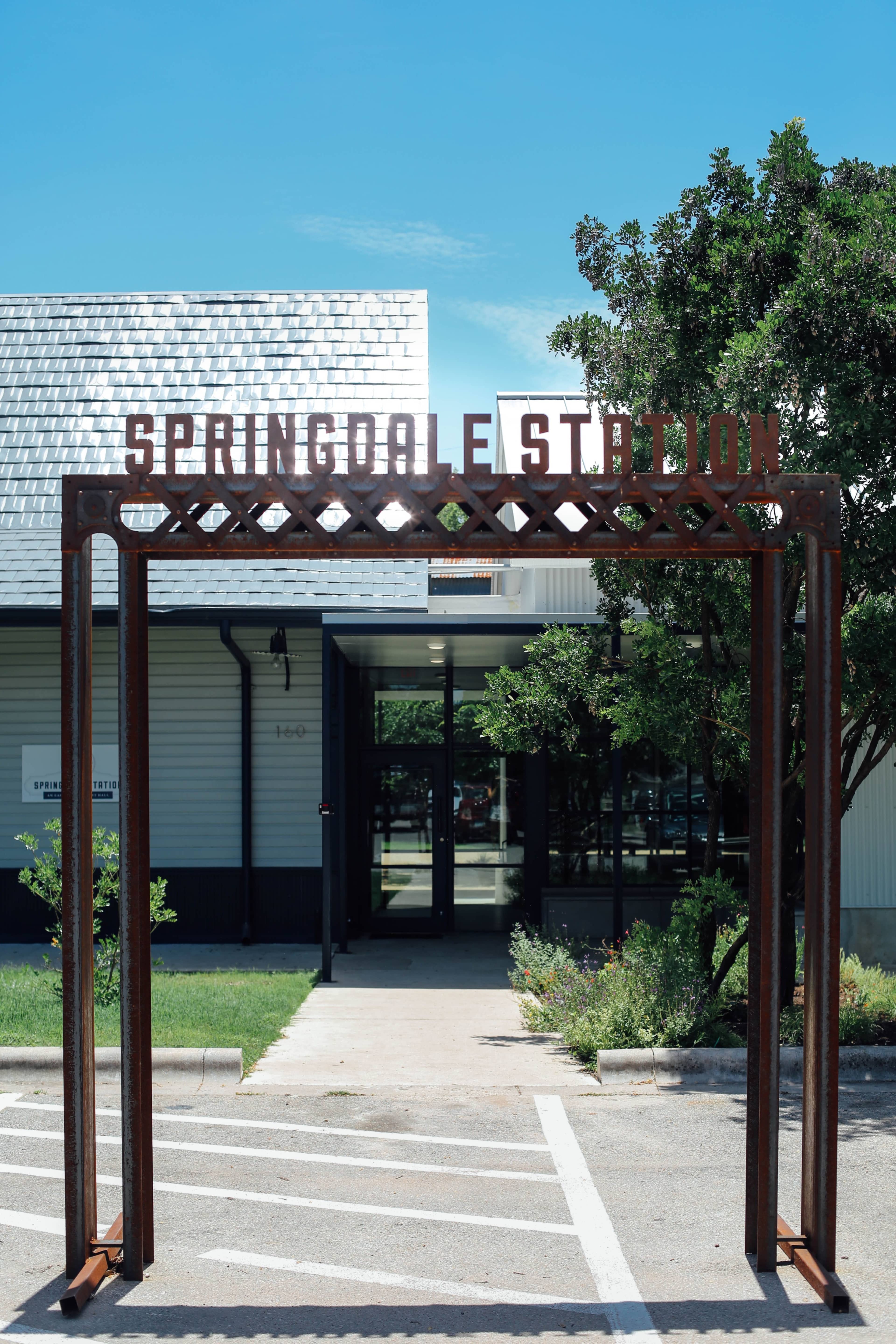 A metal archway with the words "SPRINGDALE STATION" at the entrance to a building surrounded by greenery and a clear blue sky.