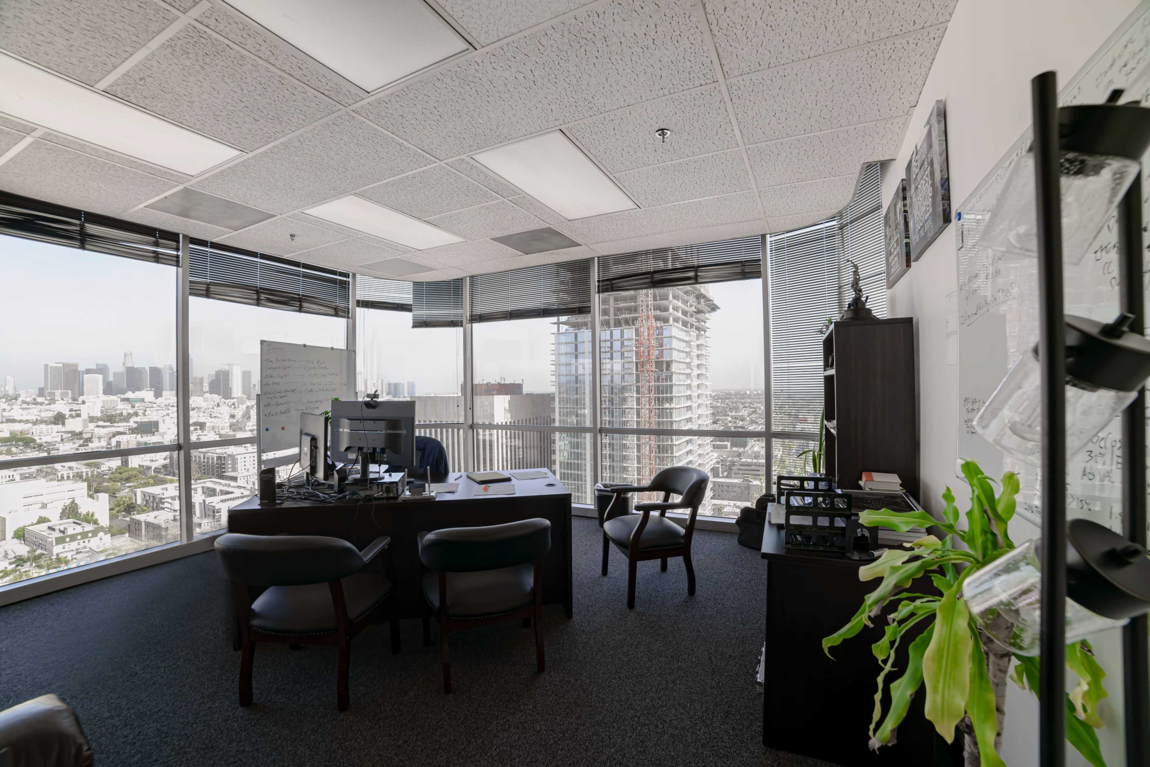 The image shows a modern office space with a large desk, several chairs, and floor-to-ceiling windows overlooking a city skyline.