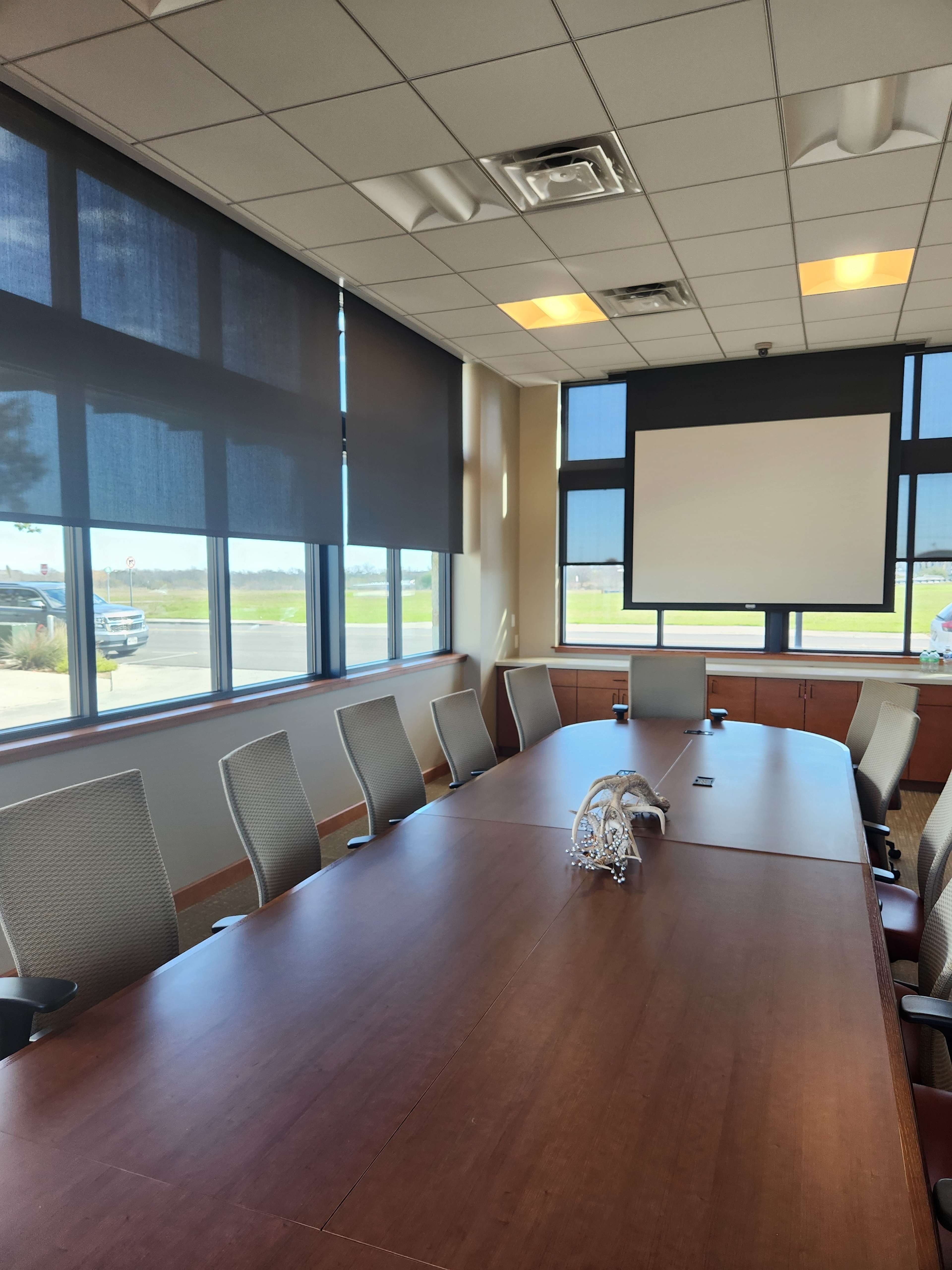 A conference room features a large wooden table surrounded by chairs, with windows allowing natural light and a projector screen at the front.