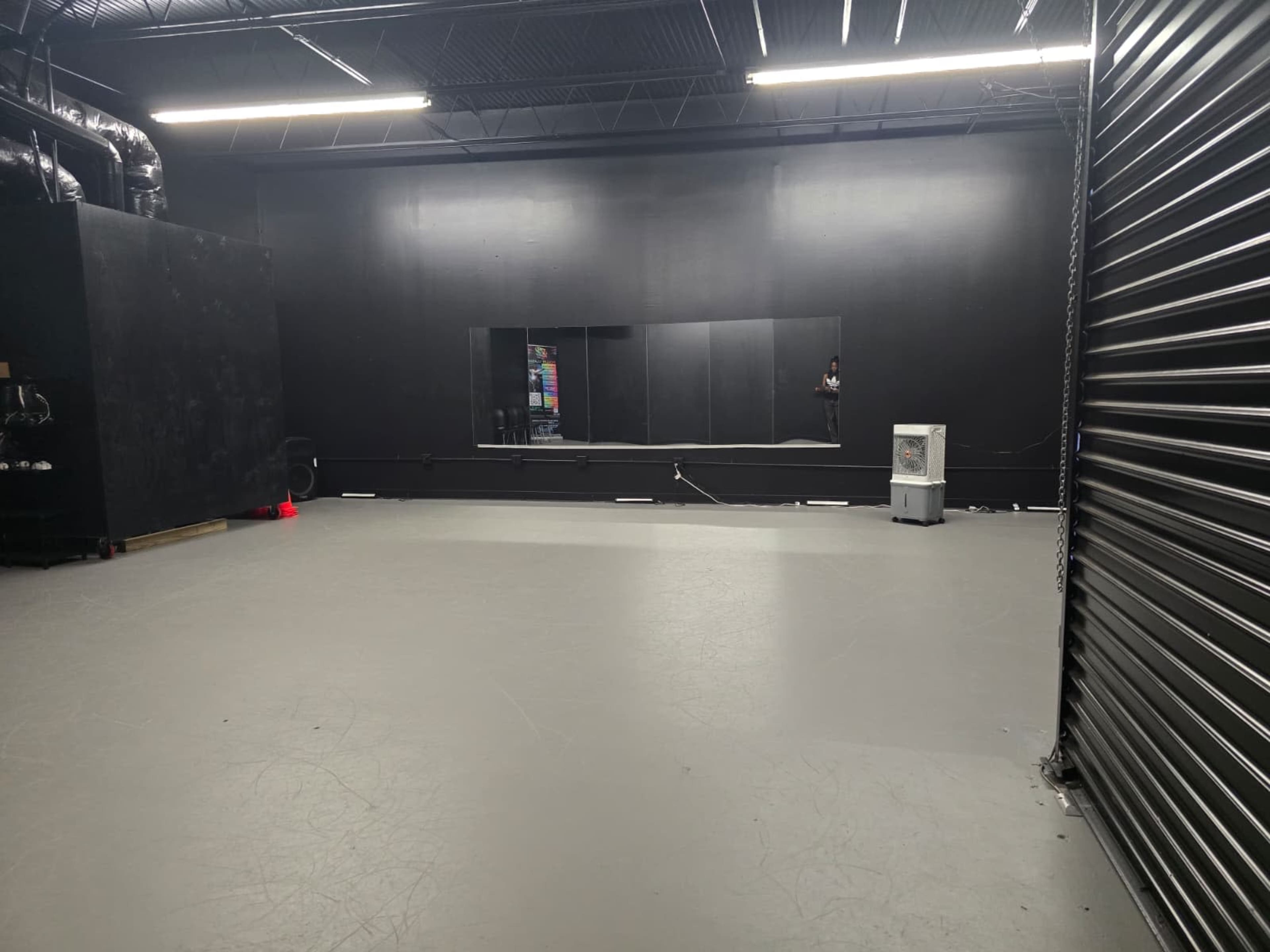 The image shows a spacious black-walled rehearsal studio with a gray floor and a large mirror along one side.