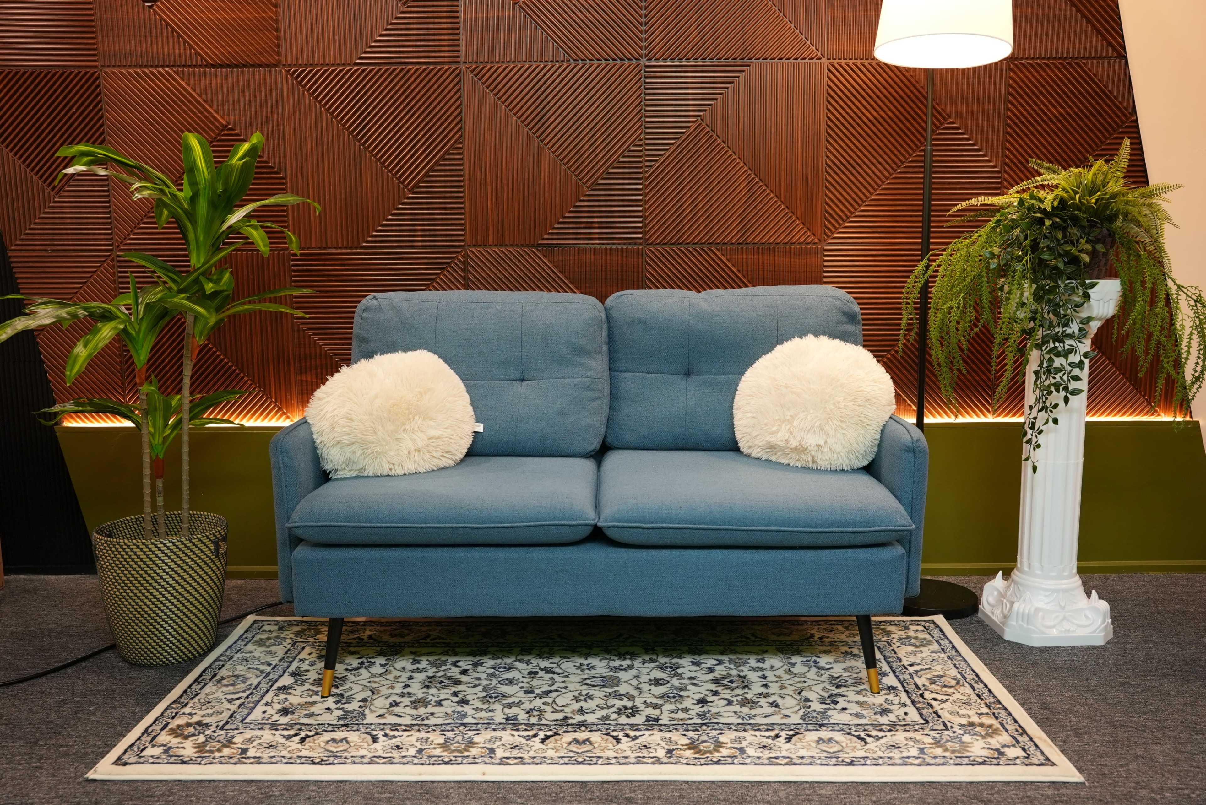 A blue sofa with two decorative pillows is positioned in front of a textured wooden wall, accompanied by a potted plant and a standing lamp.