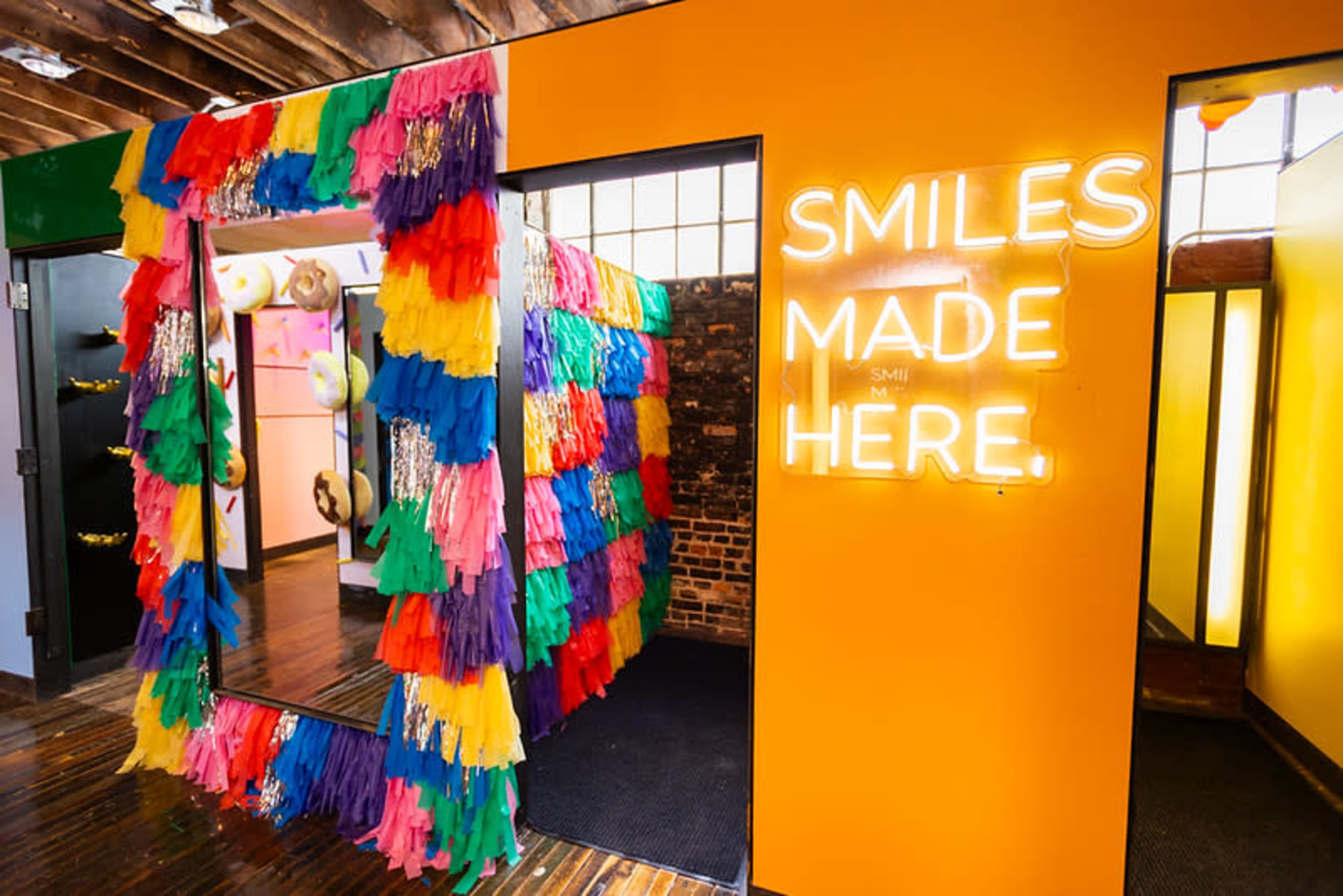 The image shows a brightly decorated entrance with a colorful fringe border and a neon sign that reads "SMILES MADE HERE."