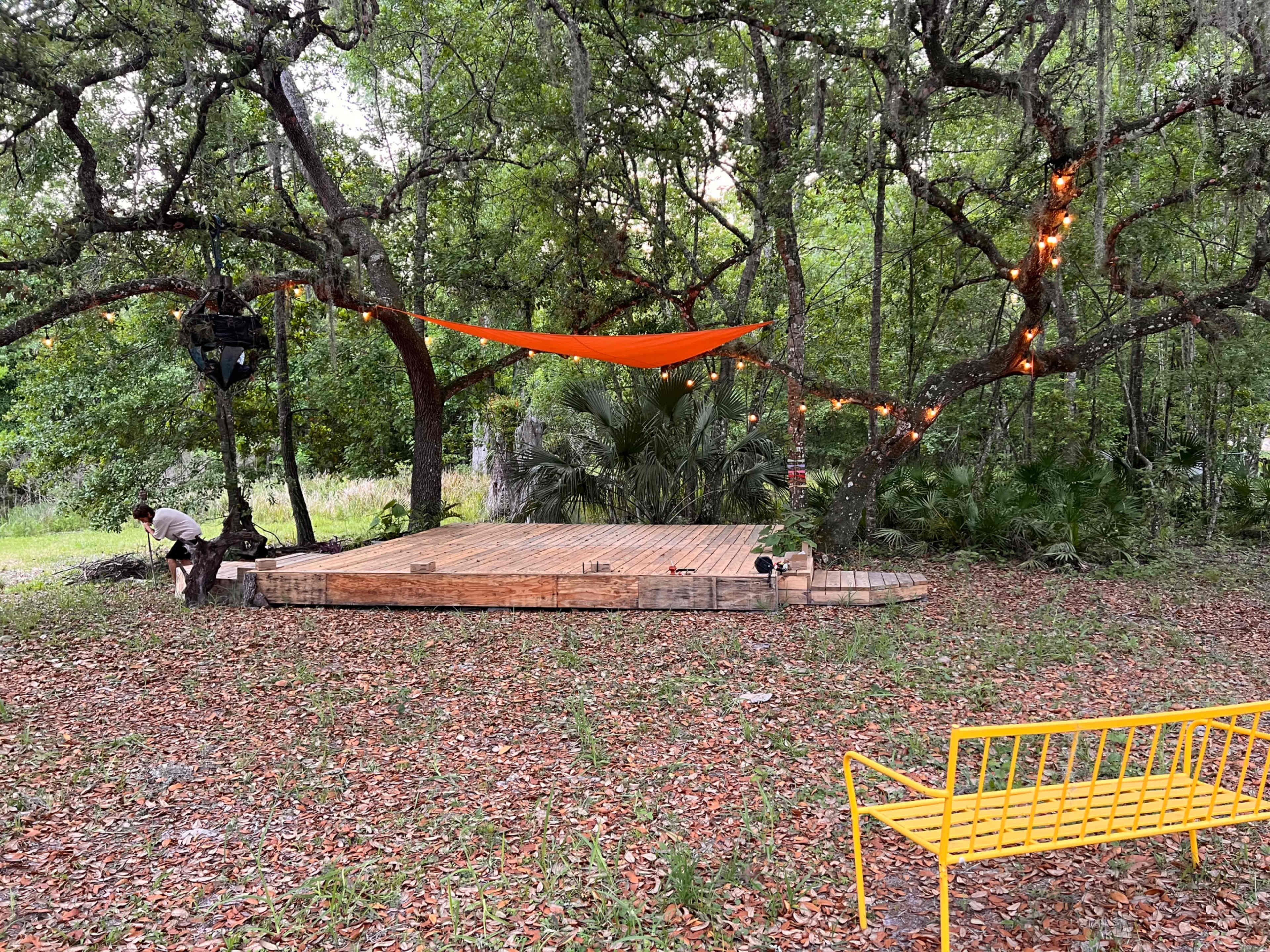 A wooden platform is set up under trees with orange fabric draped overhead and string lights, while a person works nearby.