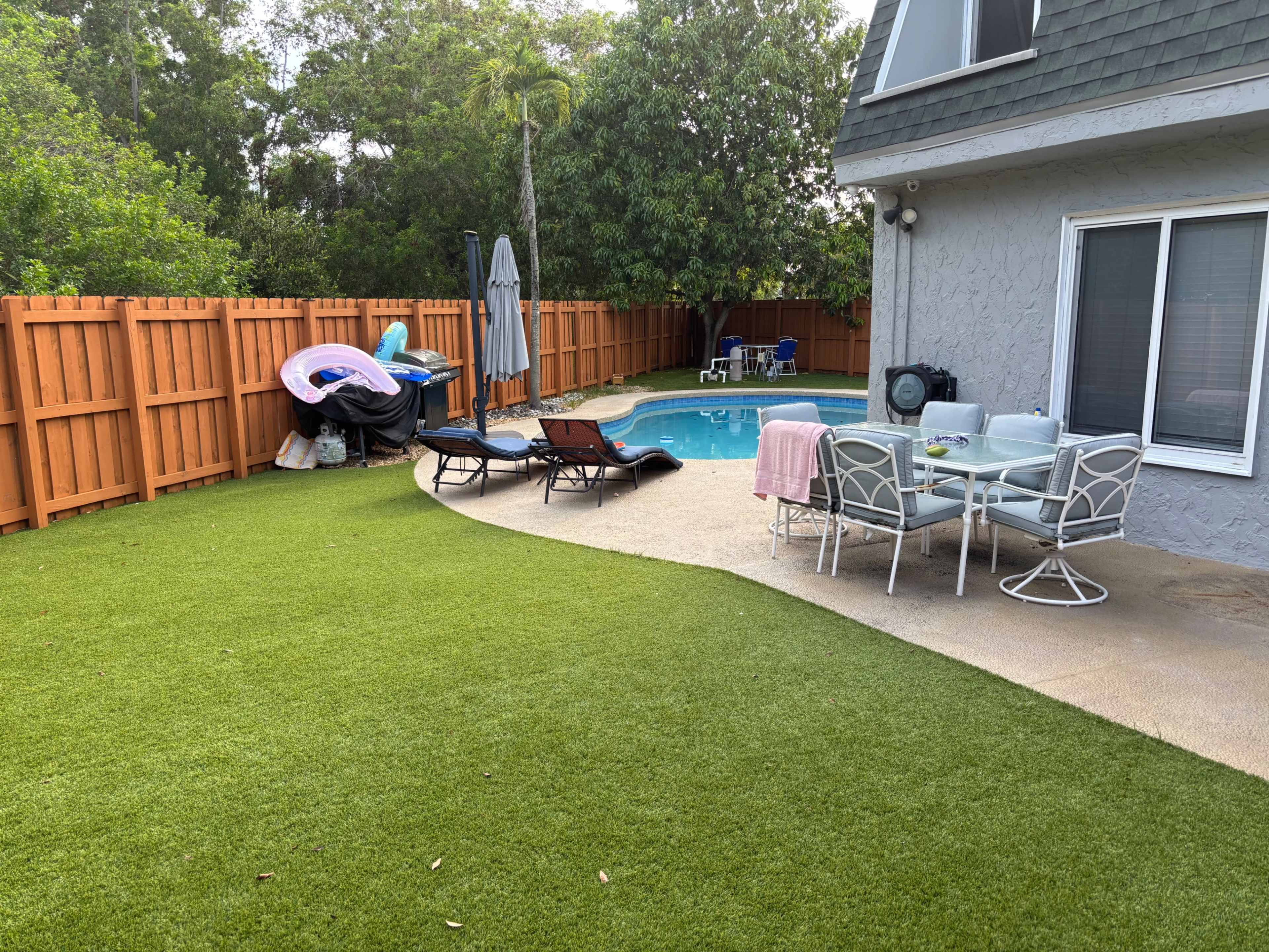 A well-maintained backyard features a swimming pool surrounded by lounge chairs, a dining set, and a wooden fence.
