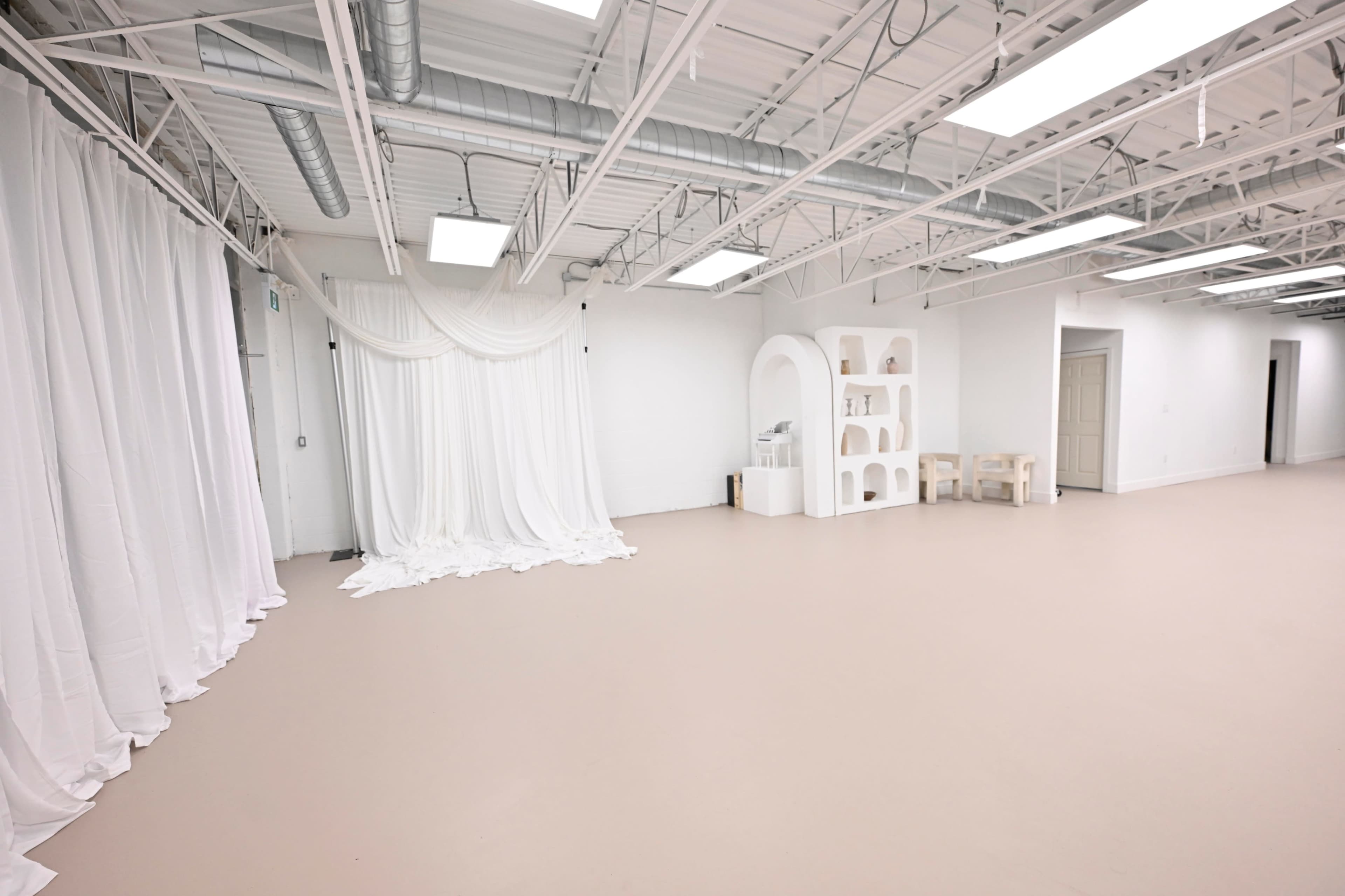 The image shows a spacious room with white walls, a light-colored floor, and a backdrop of draped white fabric on one side, alongside a minimalist shelving unit and a small table.